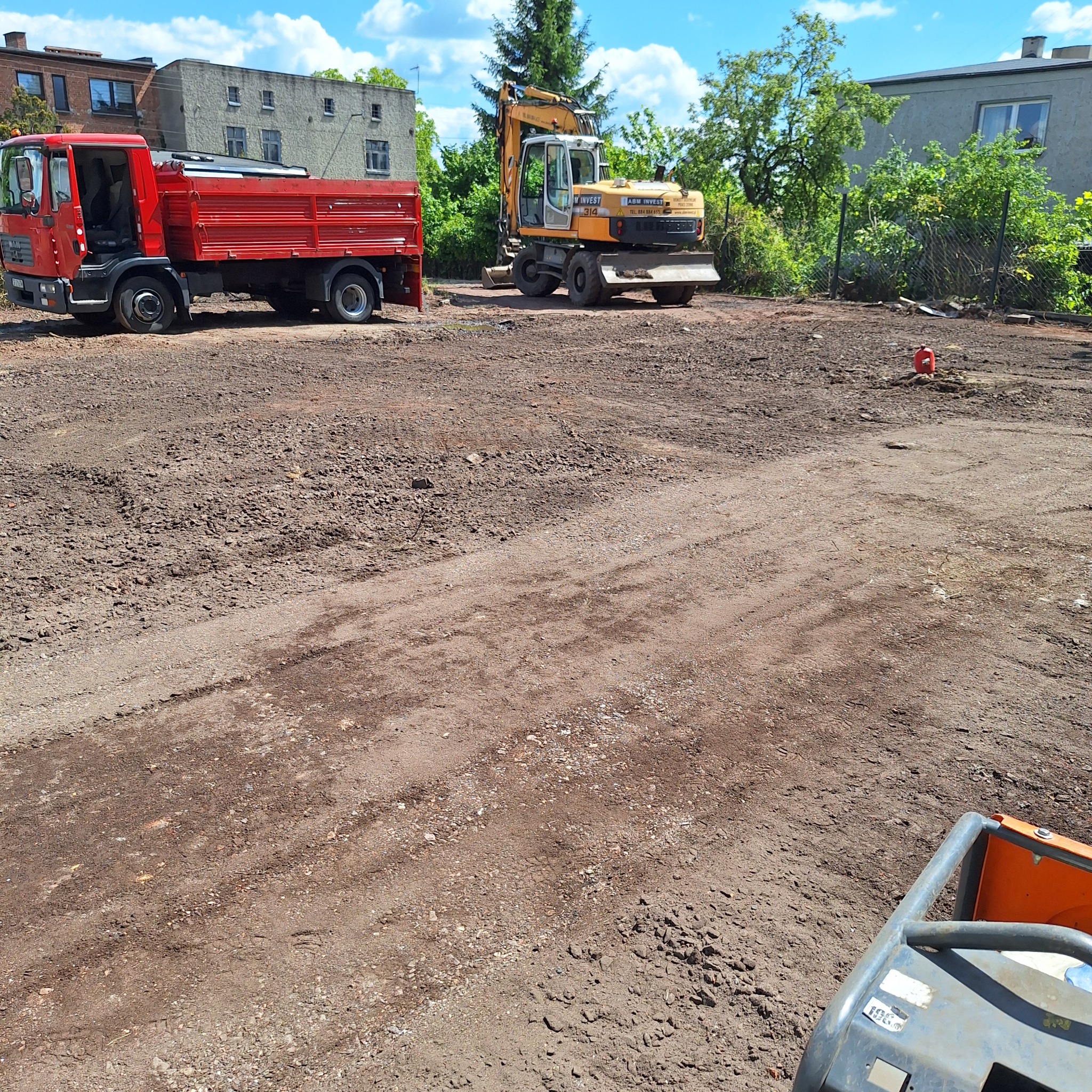 Czerwona wywrotka i żółta koparka na wyrównanym, brązowym terenie budowy. W tle budynki i drzewa pod błękitnym niebem. Częściowo widoczna zagęszczarka.