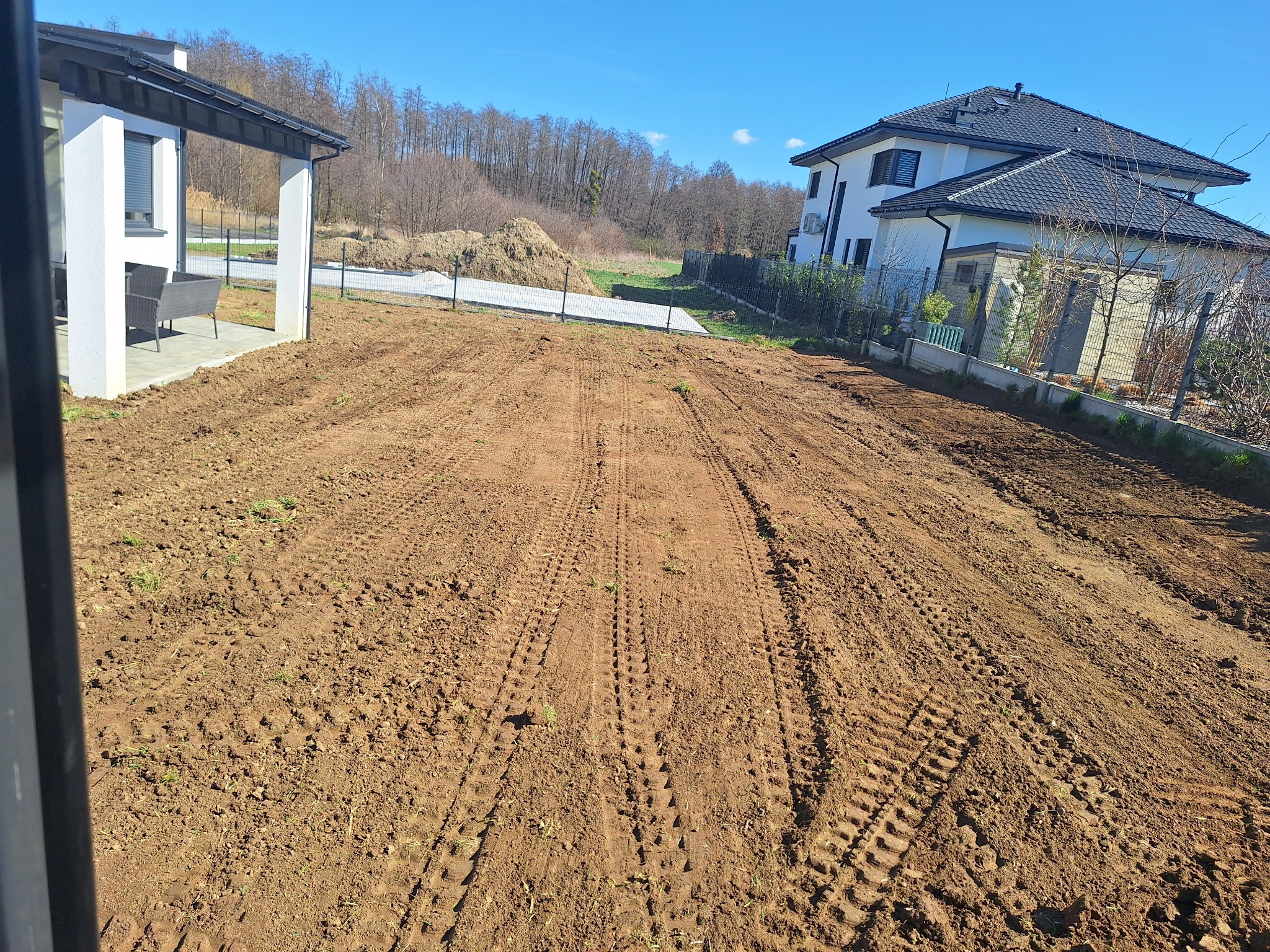 Przygotowany teren pod budowę domu jednorodzinnego z widocznymi śladami opon po maszynach budowlanych. W tle dom i drzewa.