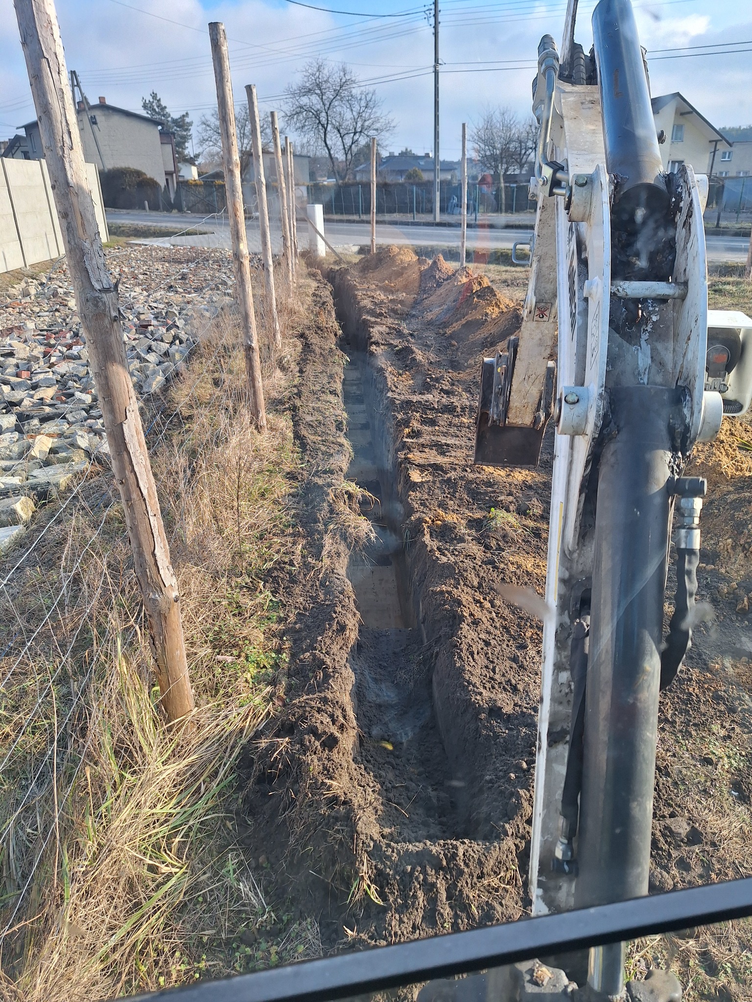 Wykop pod drenaż wykonany minikoparką w Żorach. Wąski rów w ziemi, widoczne drewniane słupki ogrodzenia i maszyna budowlana. Prace ziemne w toku.
