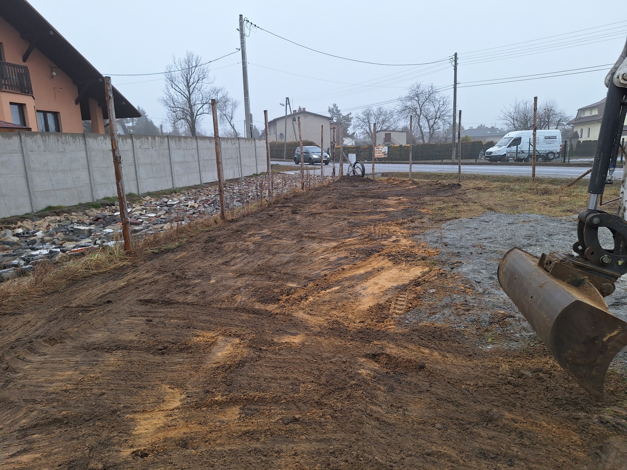Przygotowanie terenu pod budowę: wyrównana ziemia, wbite słupki, widoczny fragment łyżki koparki. W tle betonowy płot, dom i ulica z samochodami.