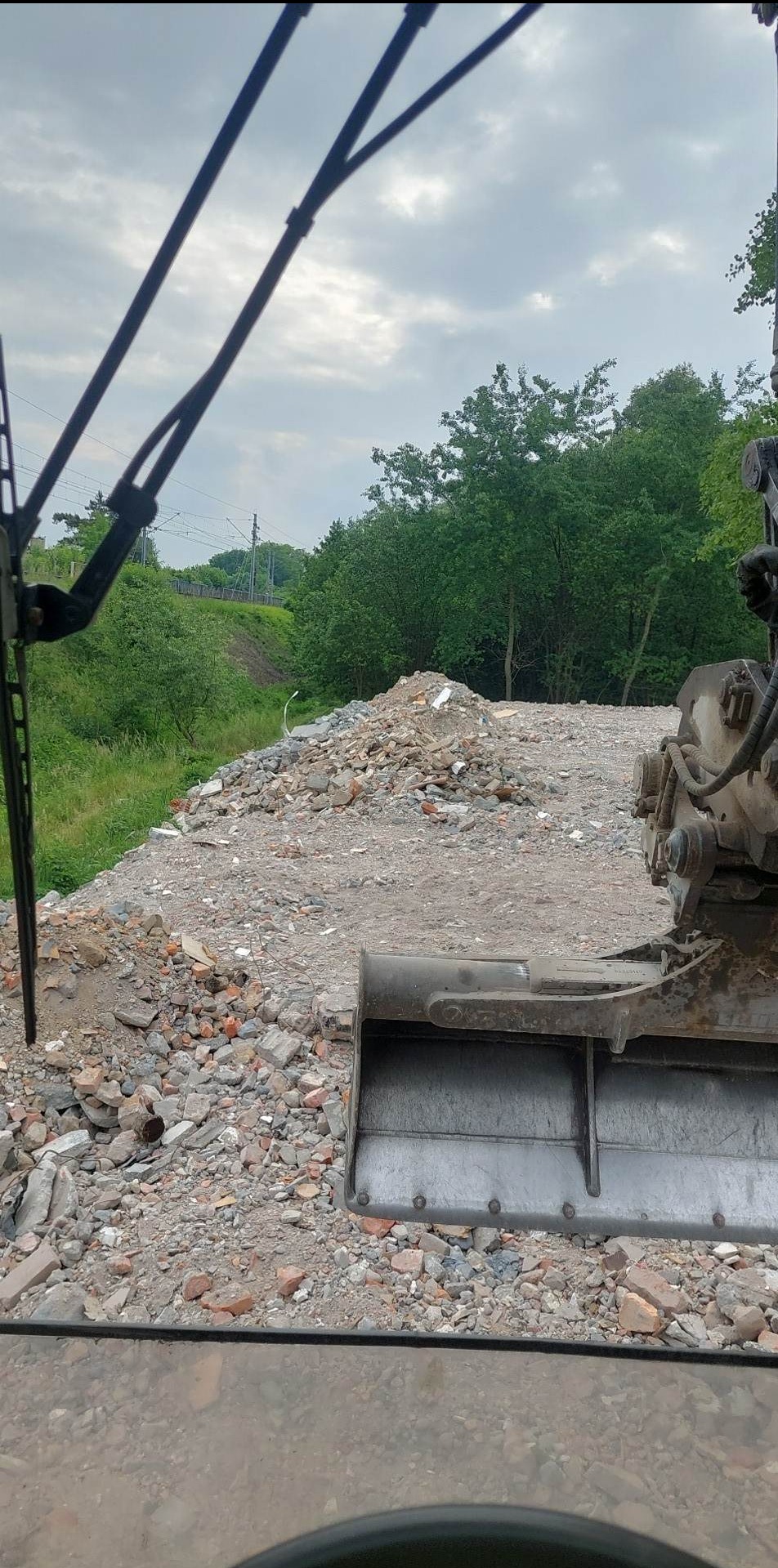 Widok z kabiny koparki na gruzowisko z fragmentami cegieł i ziemi, w tle zieleń drzew i fragment torów kolejowych. Na pierwszym planie lemiesz koparki.
