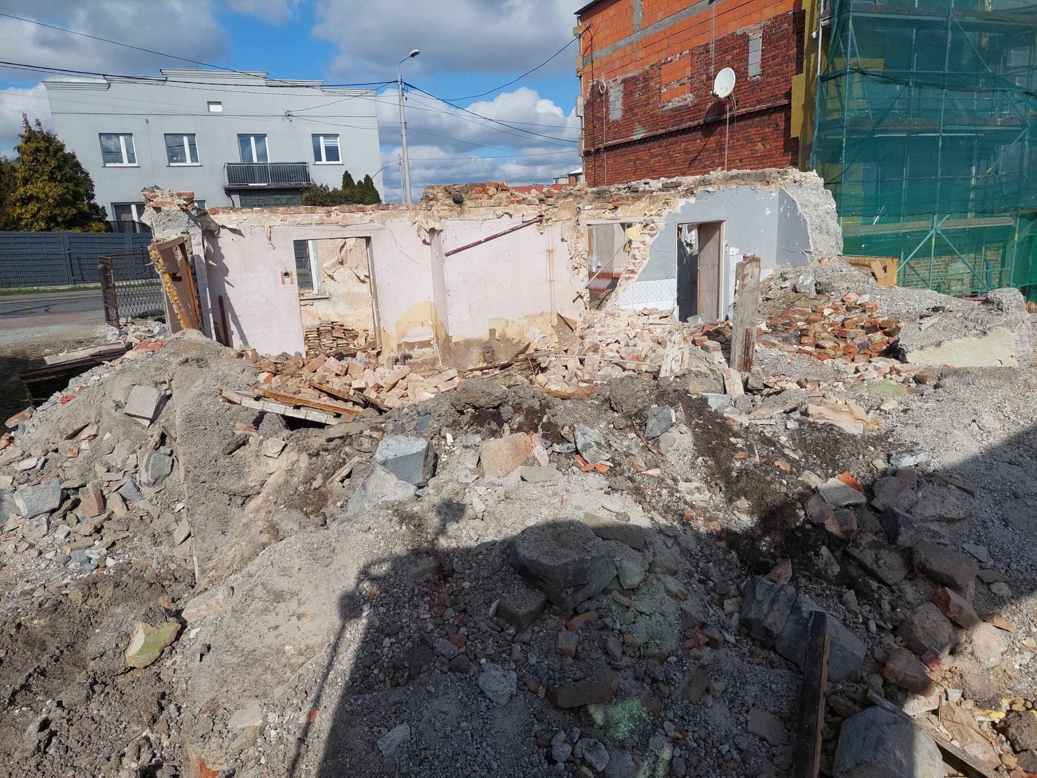 Częściowo zburzony budynek z odsłoniętymi ścianami i stertą gruzu. W tle widoczne inne budynki i błękitne niebo z chmurami. Widoczne pozostałości po oknach i drzwiach.