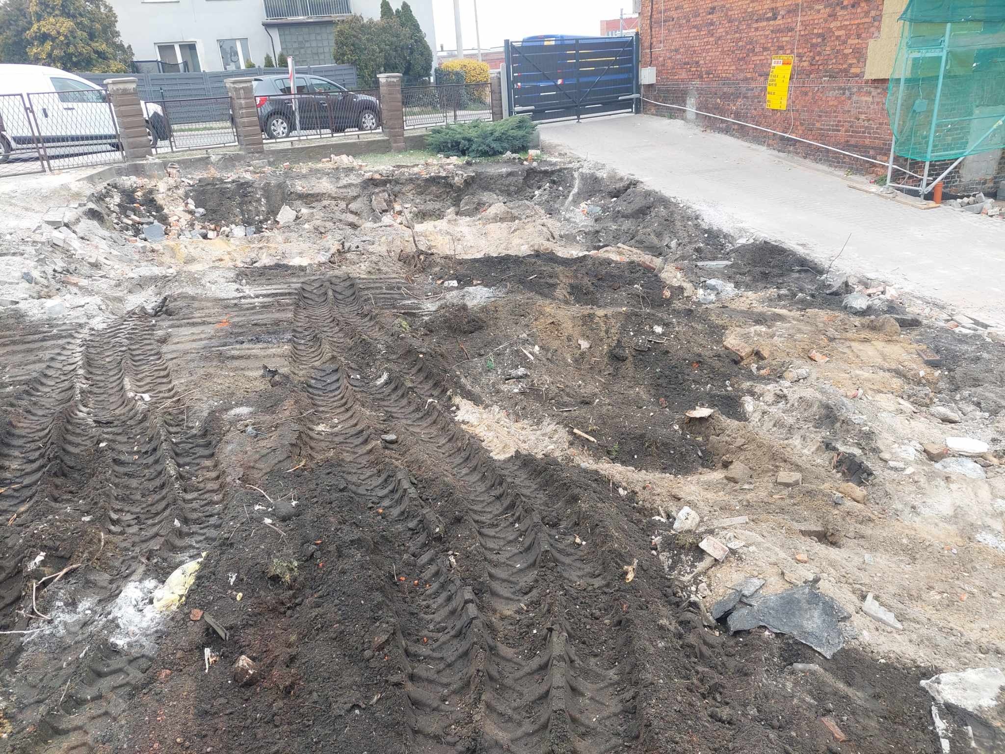 Wykonany wykop pod fundamenty z widocznymi śladami opon ciężkiego sprzętu. W tle ogrodzenie posesji i budynek z czerwonej cegły.