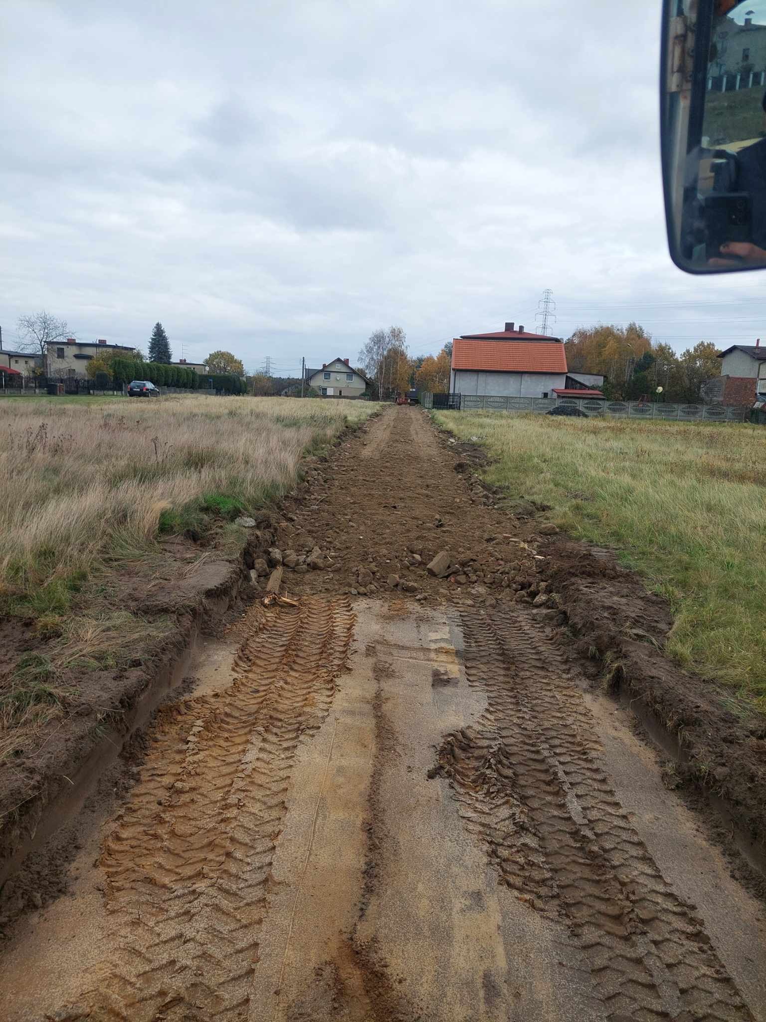 Ślady opon na błotnistej drodze gruntowej, widok z kabiny maszyny budowlanej, w tle zabudowania wiejskie pod pochmurnym niebem. Prace ziemne w toku.