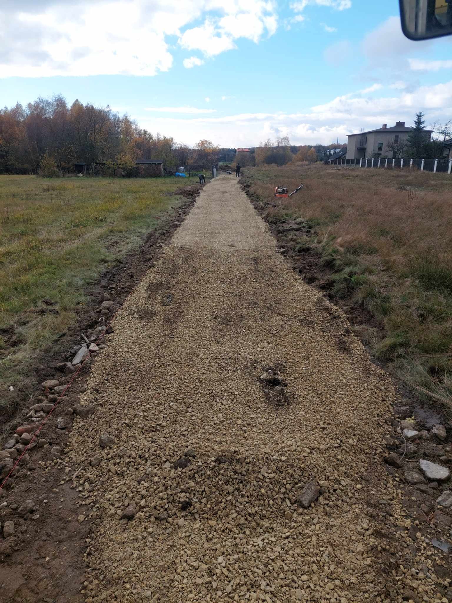 Utwardzona droga gruntowa z jasnym kruszywem, widoczne ślady po bokach, w tle drzewa i budynek. Prace ziemne w toku, zarysowany kształt drogi.