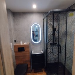 JARKO - Nowoczesna łazienka po remoncie: kabina prysznicowa z czarną ramą, podświetlane lustro owalne, drewniana podłoga i szare płytki ścienne. Widoczne elementy armatury.