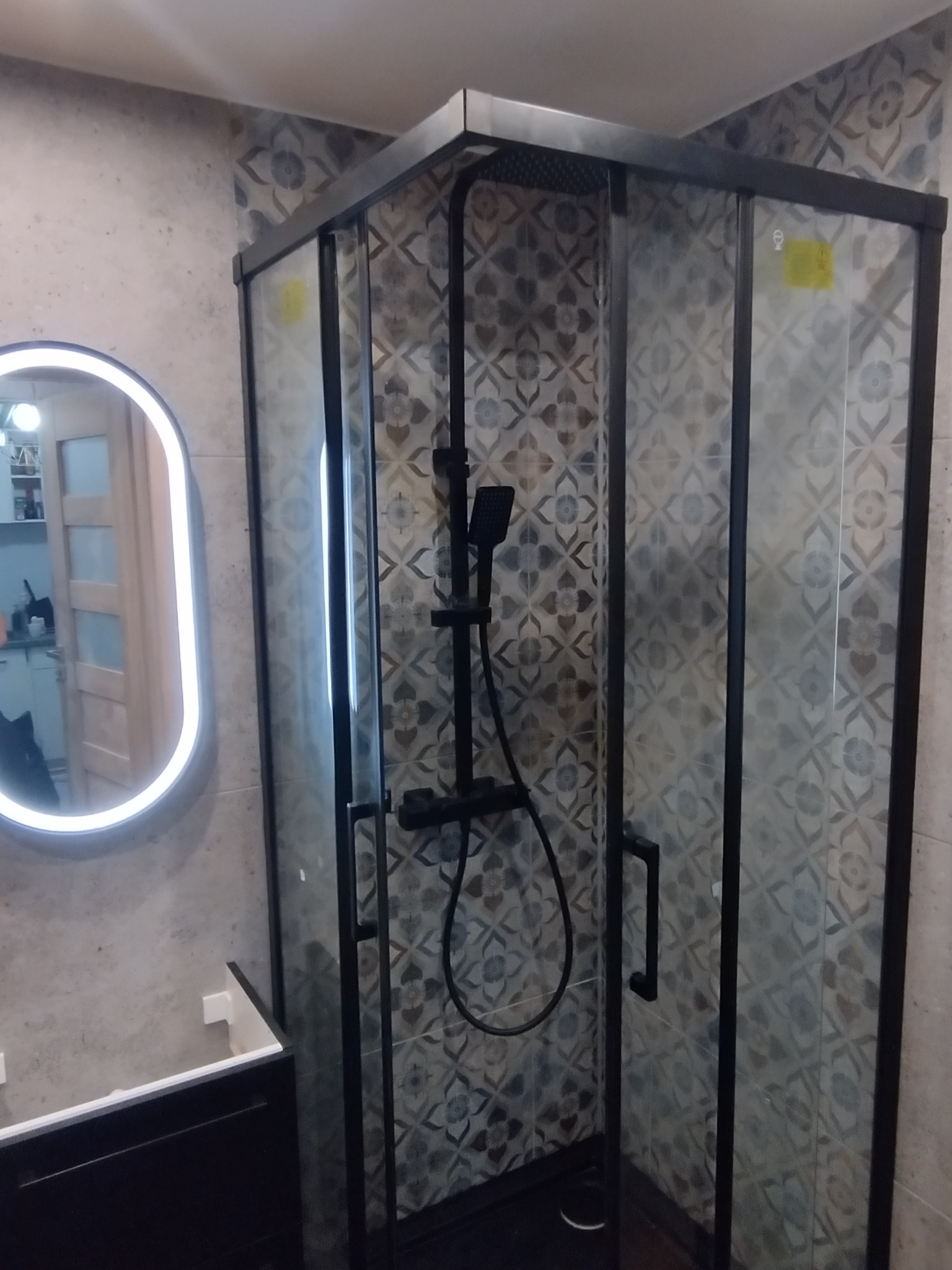 Nowoczesna kabina prysznicowa z czarnymi profilami i deszczownicą, wzorzyste płytki na ścianach, owalne lustro z podświetleniem LED. Wykończona łazienka.