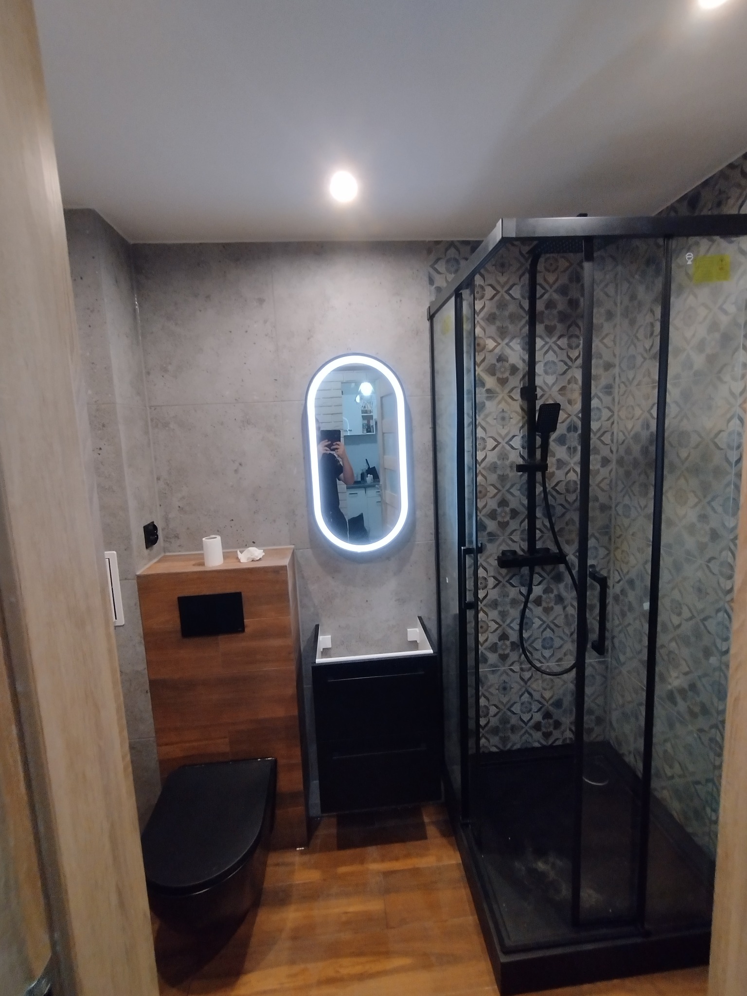 Nowoczesna łazienka po remoncie: kabina prysznicowa z czarną ramą, podświetlane lustro owalne, drewniana podłoga i szare płytki ścienne. Widoczne elementy armatury.