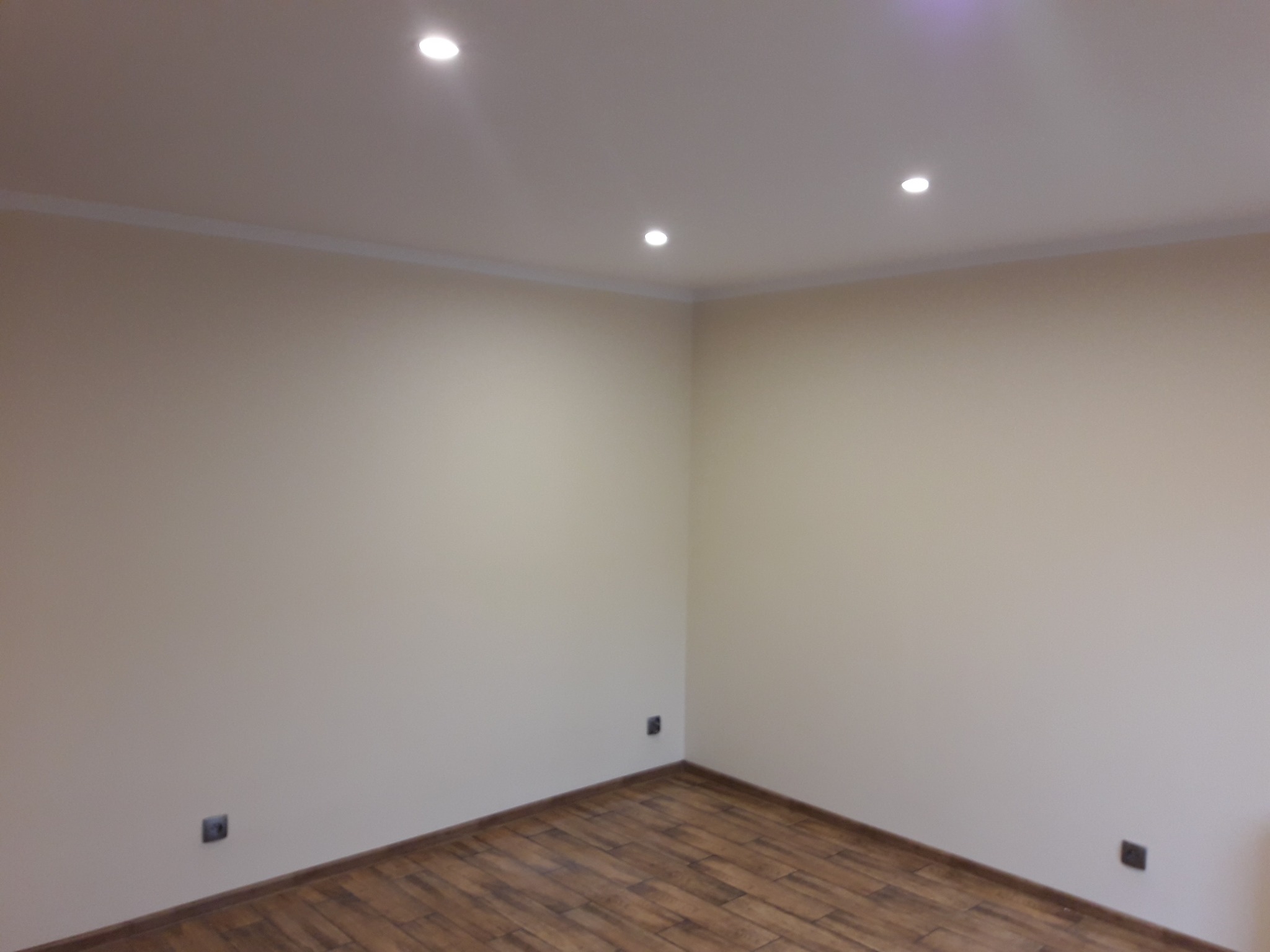 Jasne wnętrze po remoncie: drewnopodobna podłoga, kremowe ściany z listwami przysufitowymi i punktowe oświetlenie sufitowe. Widoczne gniazdka elektryczne.