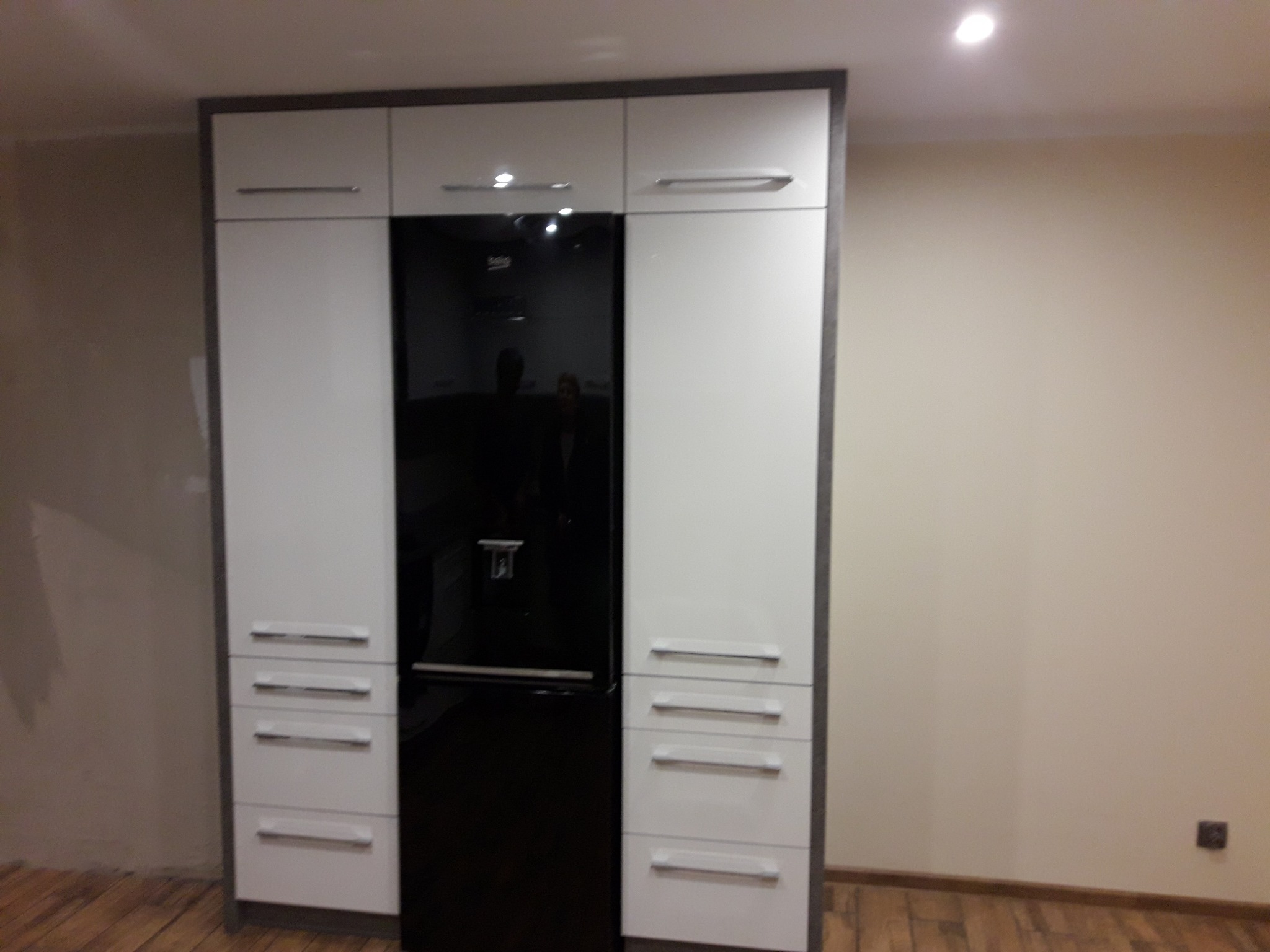 Nowoczesna zabudowa kuchenna z białymi szafkami i czarną lodówką, wykończona szarymi obramowaniami. Widok na fragment drewnianej podłogi i beżową ścianę.