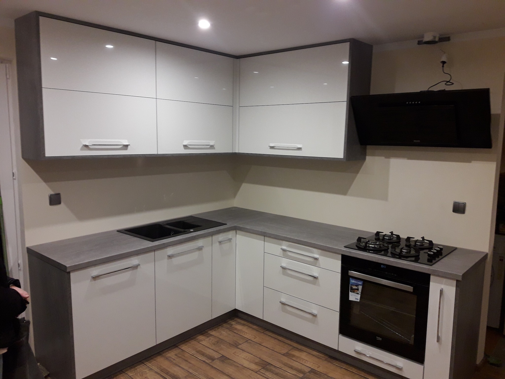 Nowoczesna kuchnia w bieli i szarości: lakierowane fronty szafek, szary blat, płyta indukcyjna i piekarnik. Drewniana podłoga i czarny okap.