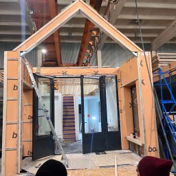 Plamchyk - Montaż modułowej konstrukcji domu z płyt warstwowych w hali produkcyjnej. Widoczne elementy konstrukcyjne, okna i drabina. W tle suwnica i oświetlenie przemysłowe.