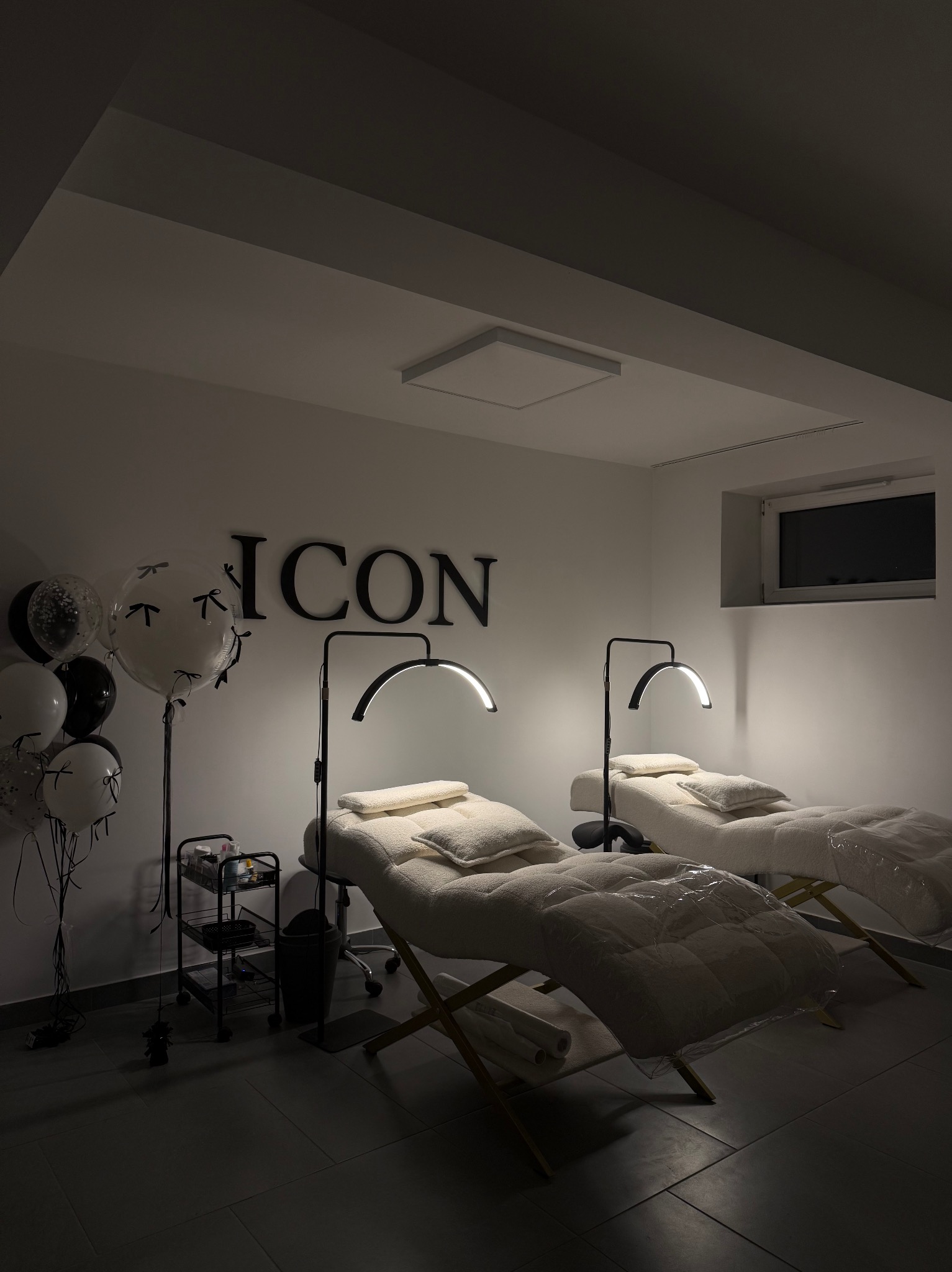 Nowoczesne wnętrze salonu kosmetycznego z dwoma fotelami zabiegowymi pod lampami, dekorowane balonami i napisem 'ICON' na ścianie. Stonowana kolorystyka.