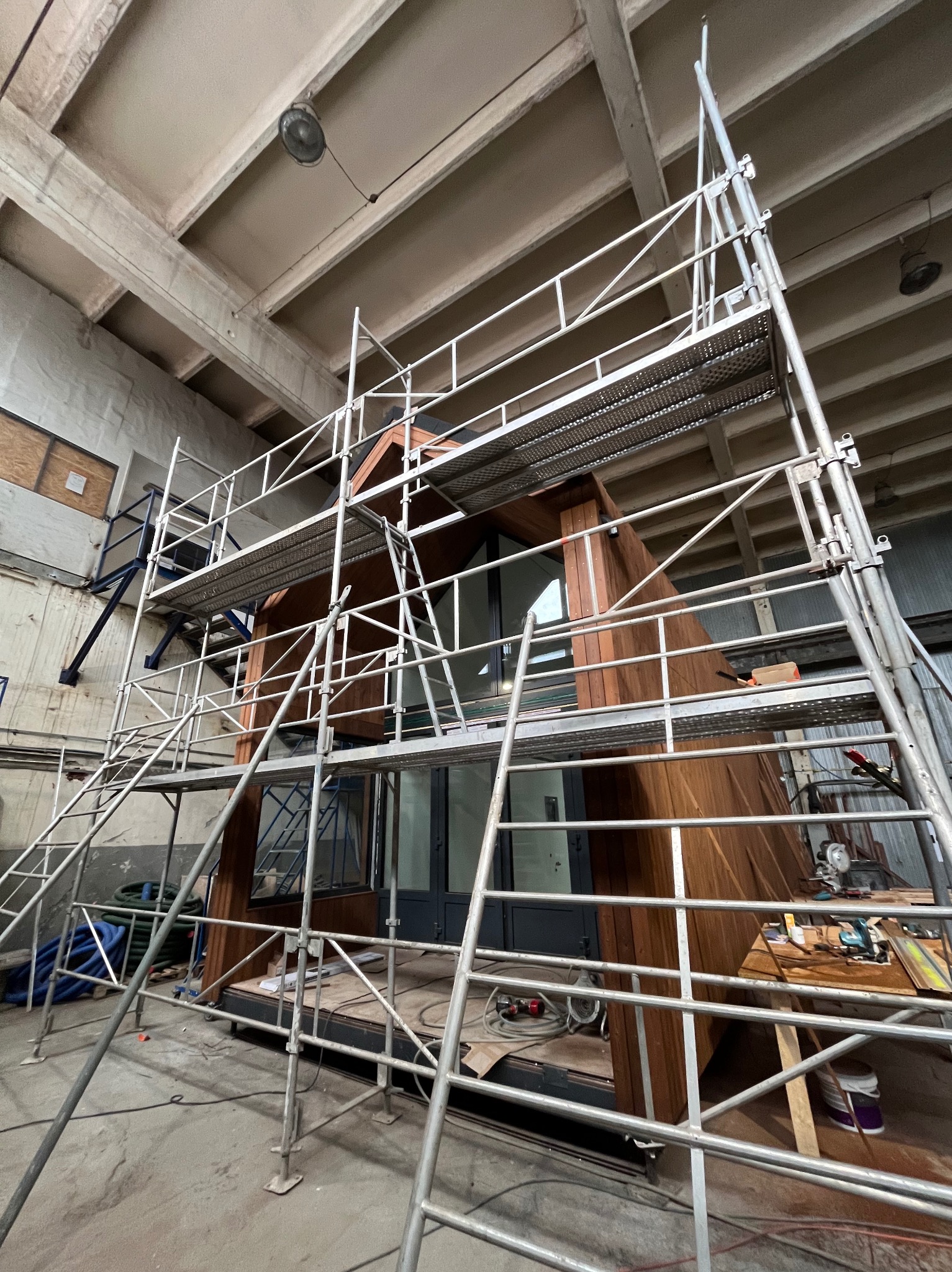 Montaż drewnianej elewacji budynku w hali, widoczne rusztowania i narzędzia. Ujęcie z dołu, surowy industrialny klimat wnętrza.