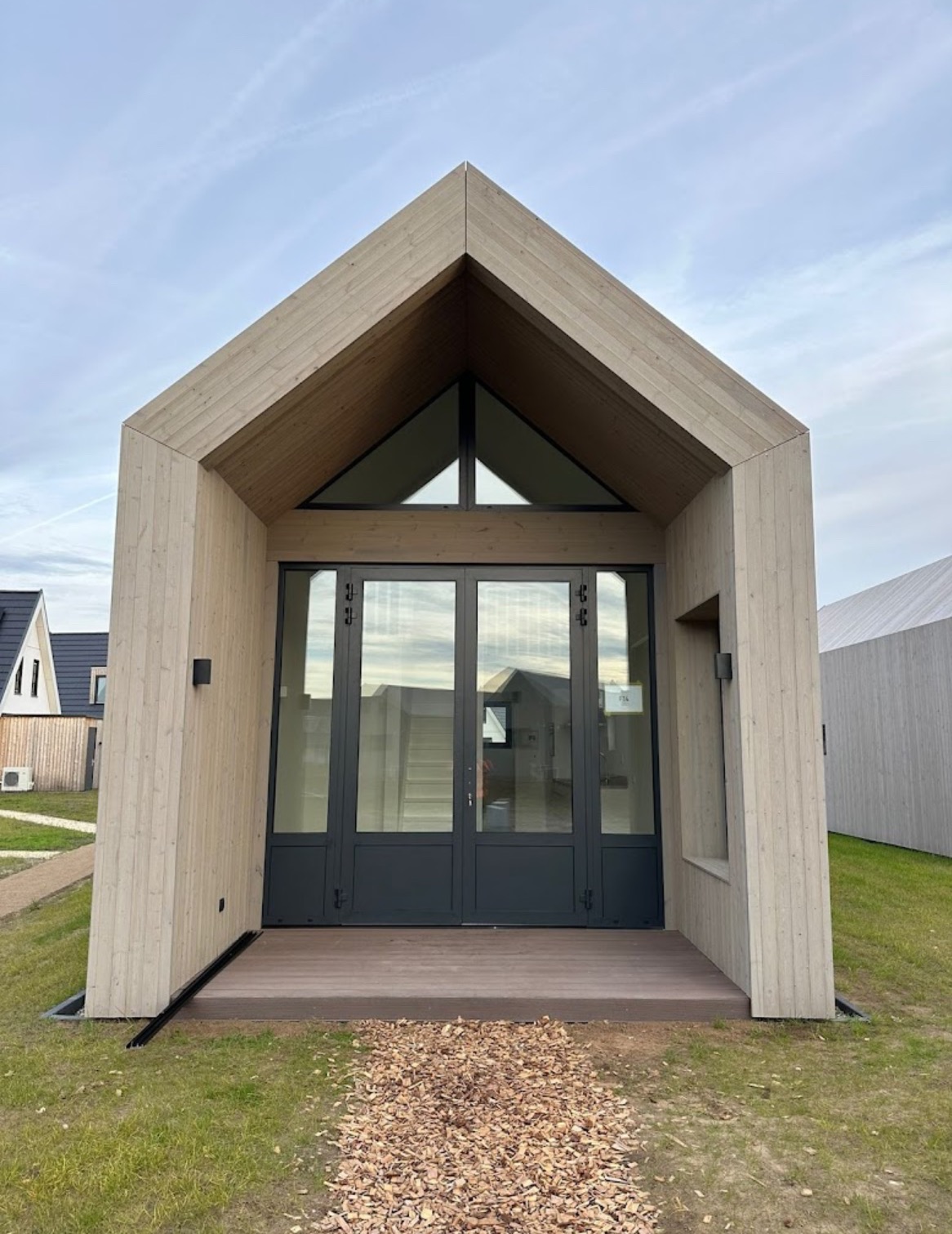 Nowoczesna elewacja domu z jasnego drewna z dużymi, szklanymi drzwiami i trójkątnym oknem nad wejściem. Minimalistyczny design, otoczenie zielone.