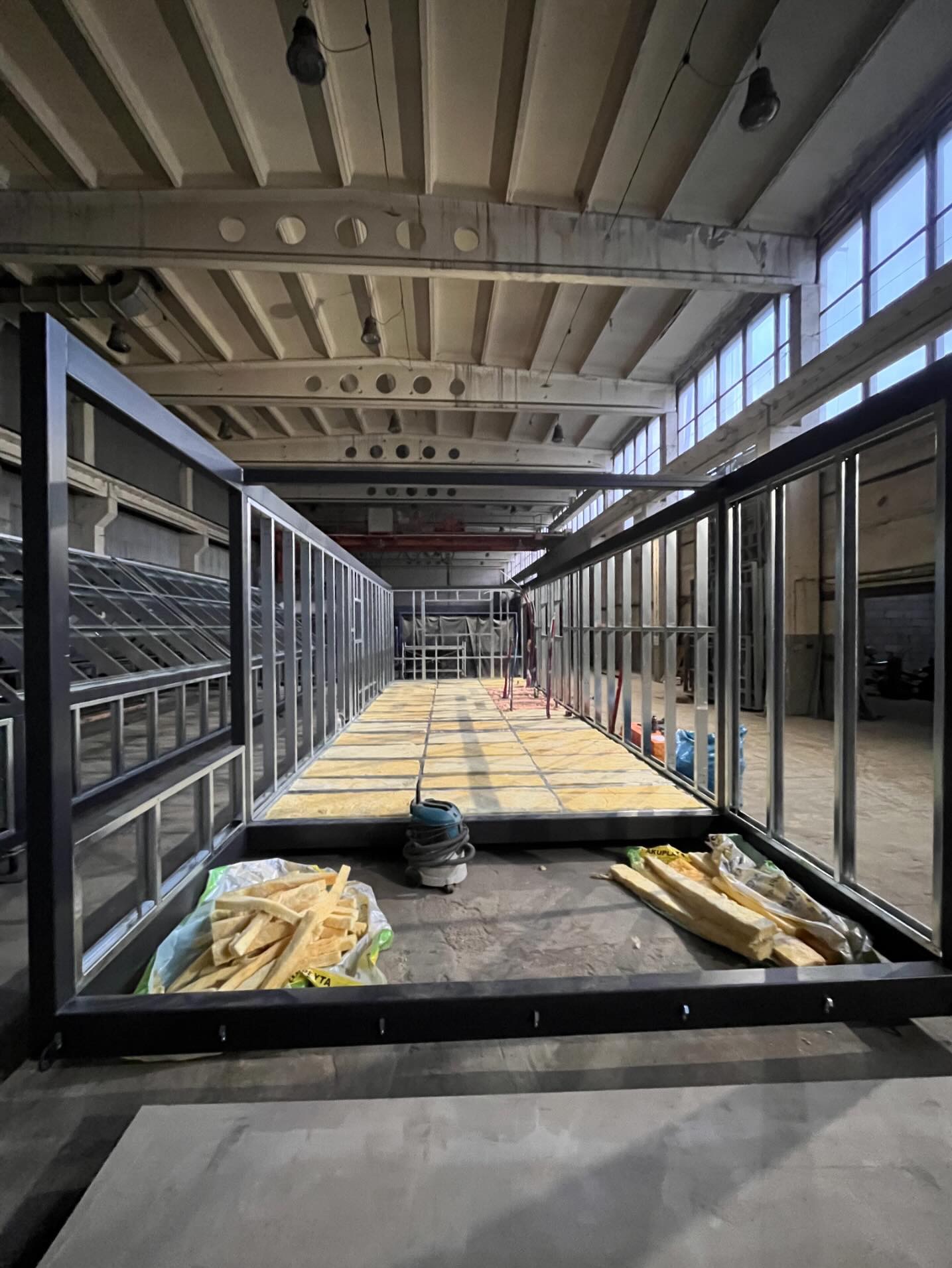 Metalowa konstrukcja szkieletowa w hali, przygotowana do ocieplenia. Widoczne materiały izolacyjne i narzędzia. Surowy, industrialny charakter wnętrza.