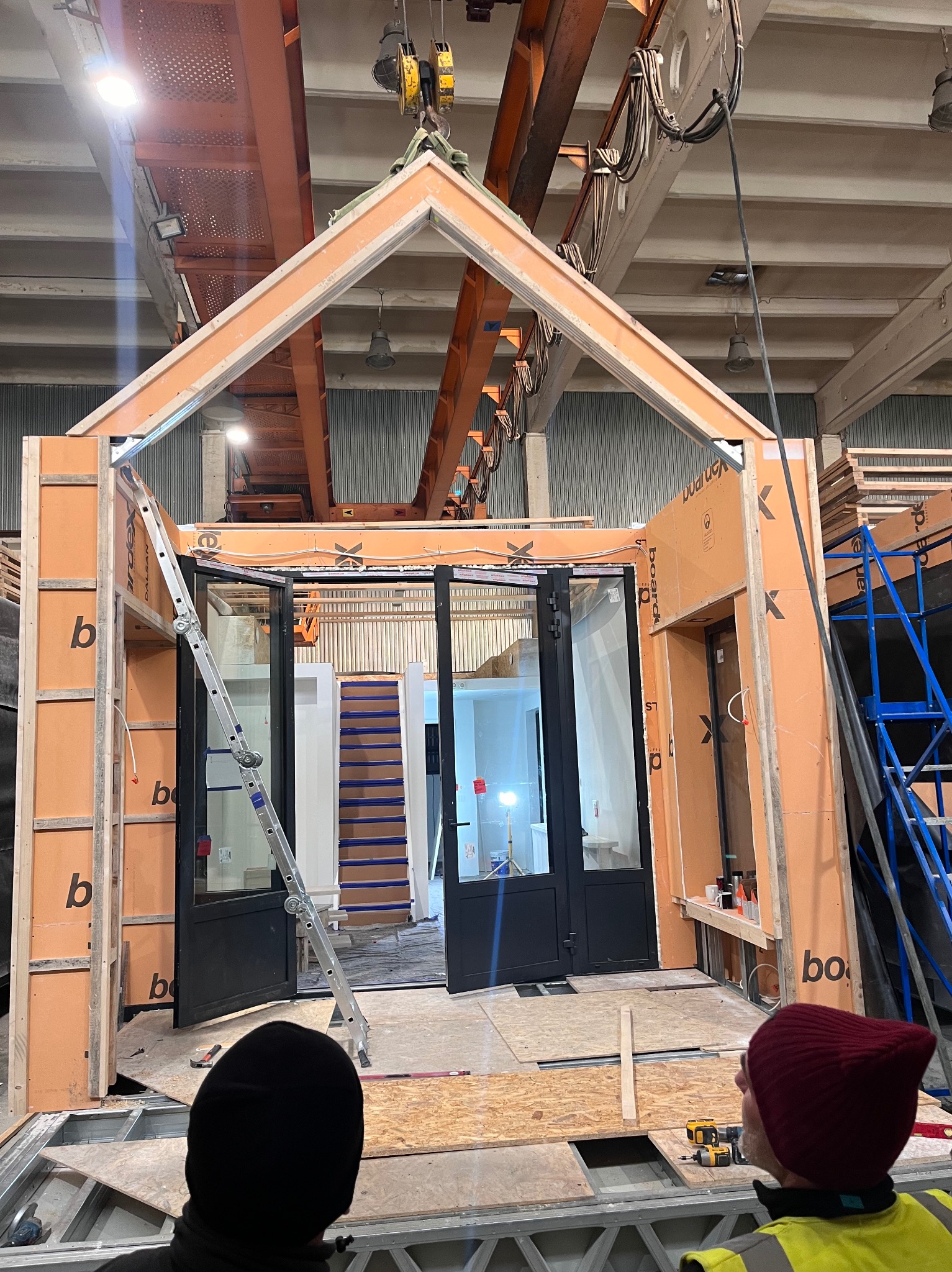 Montaż modułowej konstrukcji domu z płyt warstwowych w hali produkcyjnej. Widoczne elementy konstrukcyjne, okna i drabina. W tle suwnica i oświetlenie przemysłowe.