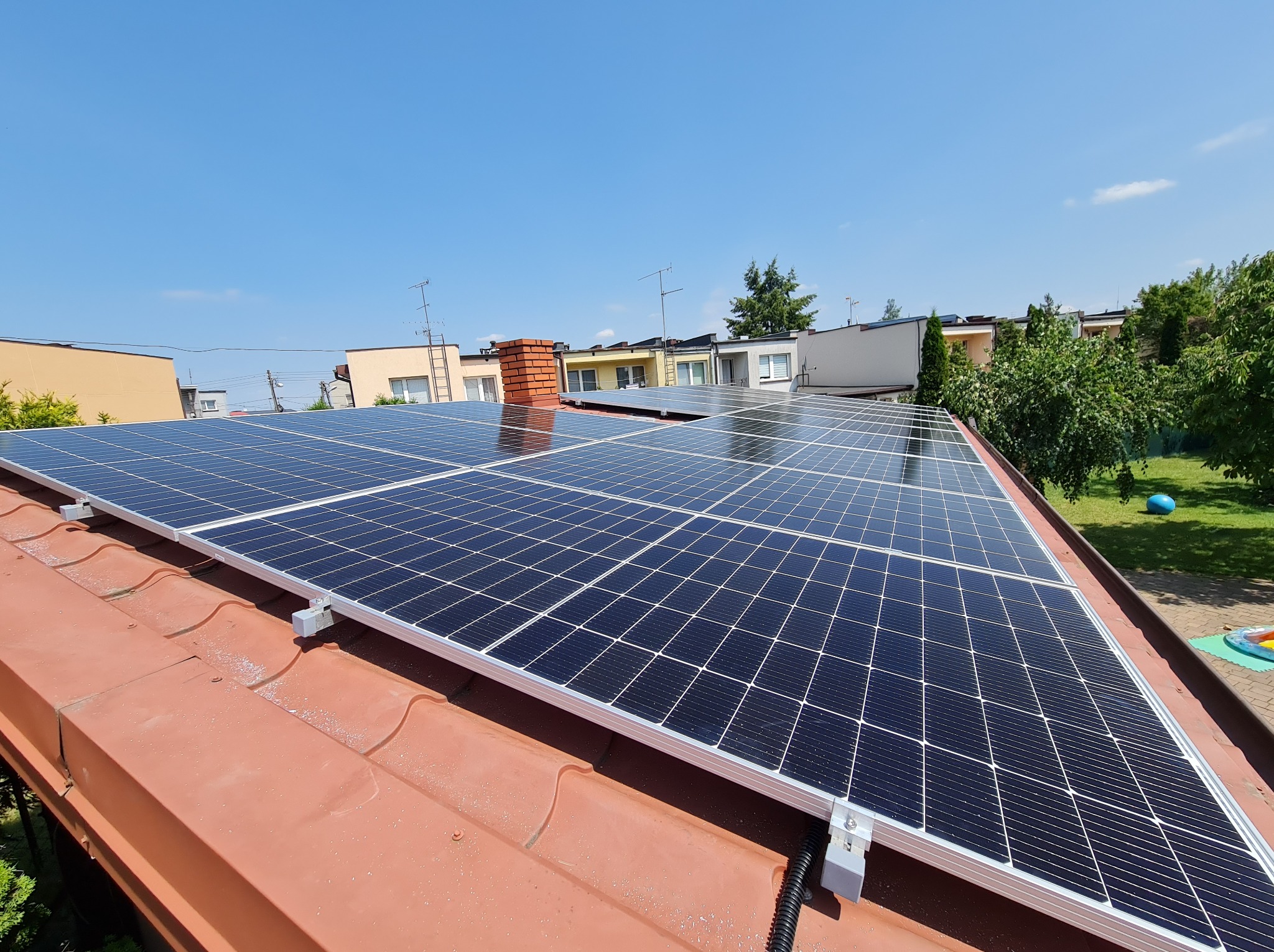 Panele fotowoltaiczne na dachu domu jednorodzinnego, widok z góry, błękitne niebo w tle. Czerwona dachówka i nowoczesne mocowania paneli.