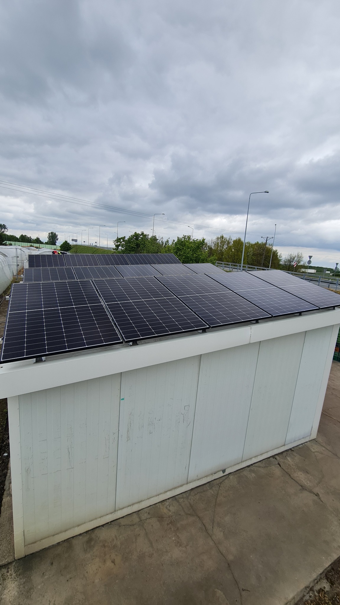 Instalacja paneli słonecznych na białym budynku z widokiem na pochmurne niebo i drzewa w tle. Panele ułożone w rzędach, równomiernie rozmieszczone na dachu.