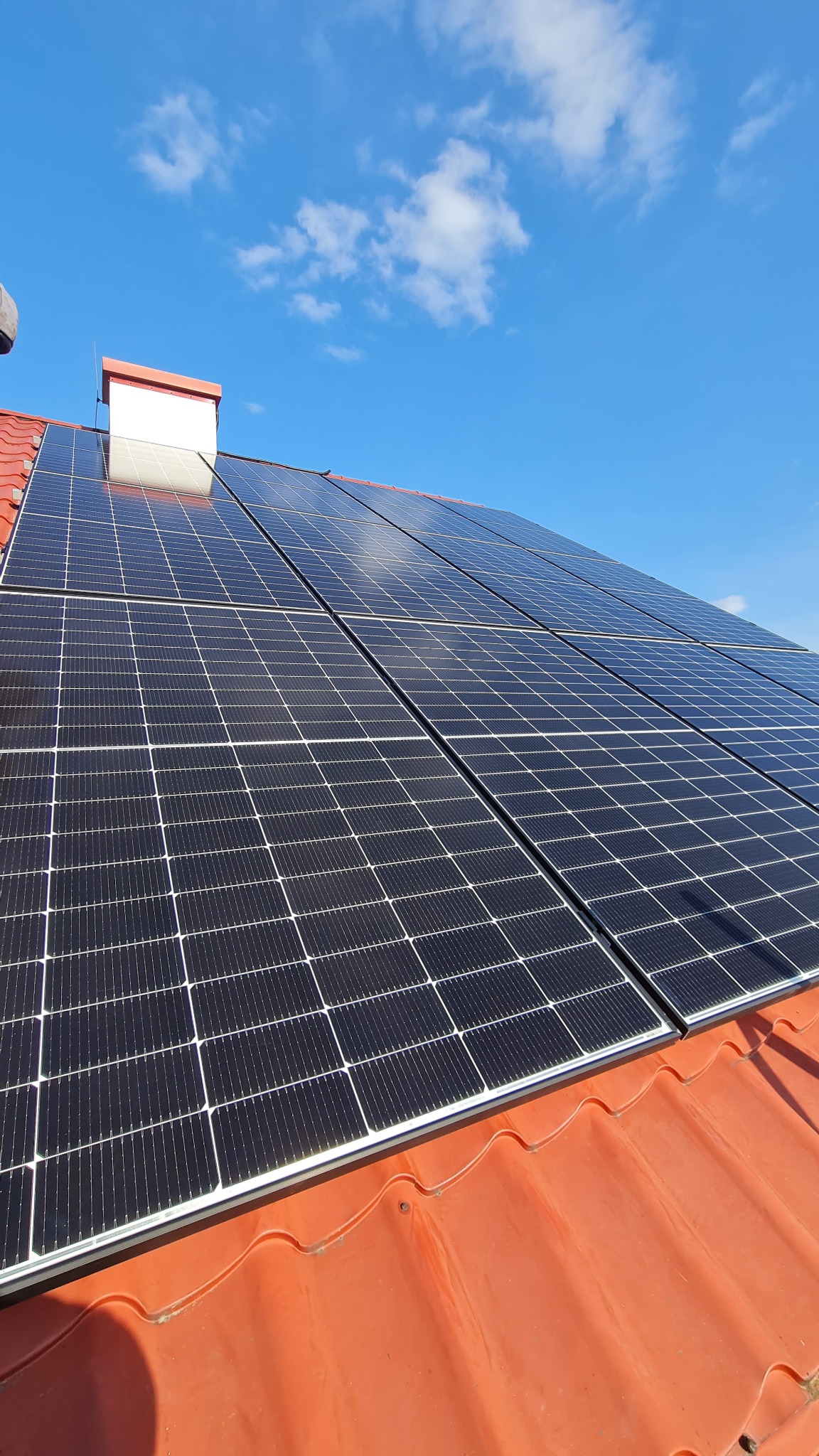 Panele słoneczne na czerwonym dachu domu w słoneczny dzień. Widoczny komin i błękitne niebo z chmurami. Instalacja fotowoltaiczna na tle nieba.