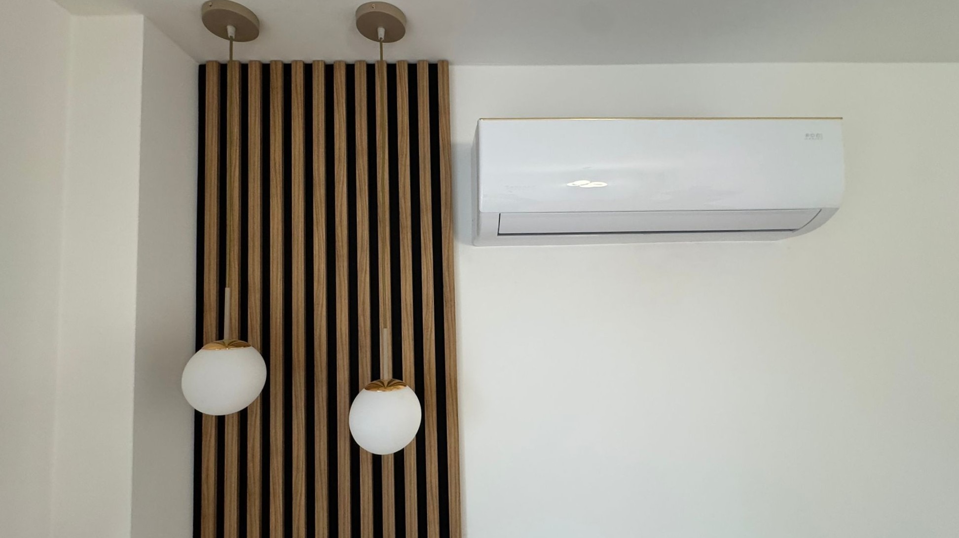 Biały klimatyzator z delikatnym złotym paskiem, zamontowany na białej ścianie obok dekoracyjnej drewnianej ścianki z lampami w kształcie kuli.