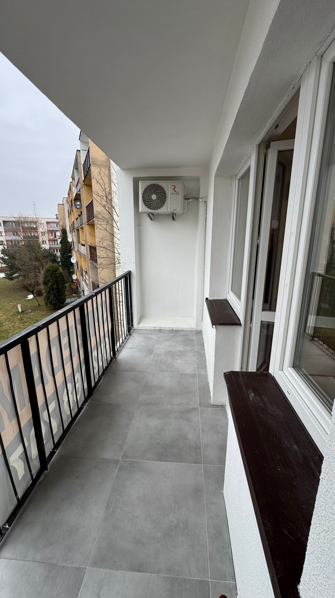 Wyremontowany balkon z szarymi płytkami, białą balustradą i zamontowanym klimatyzatorem. Widok na budynek mieszkalny w tle.