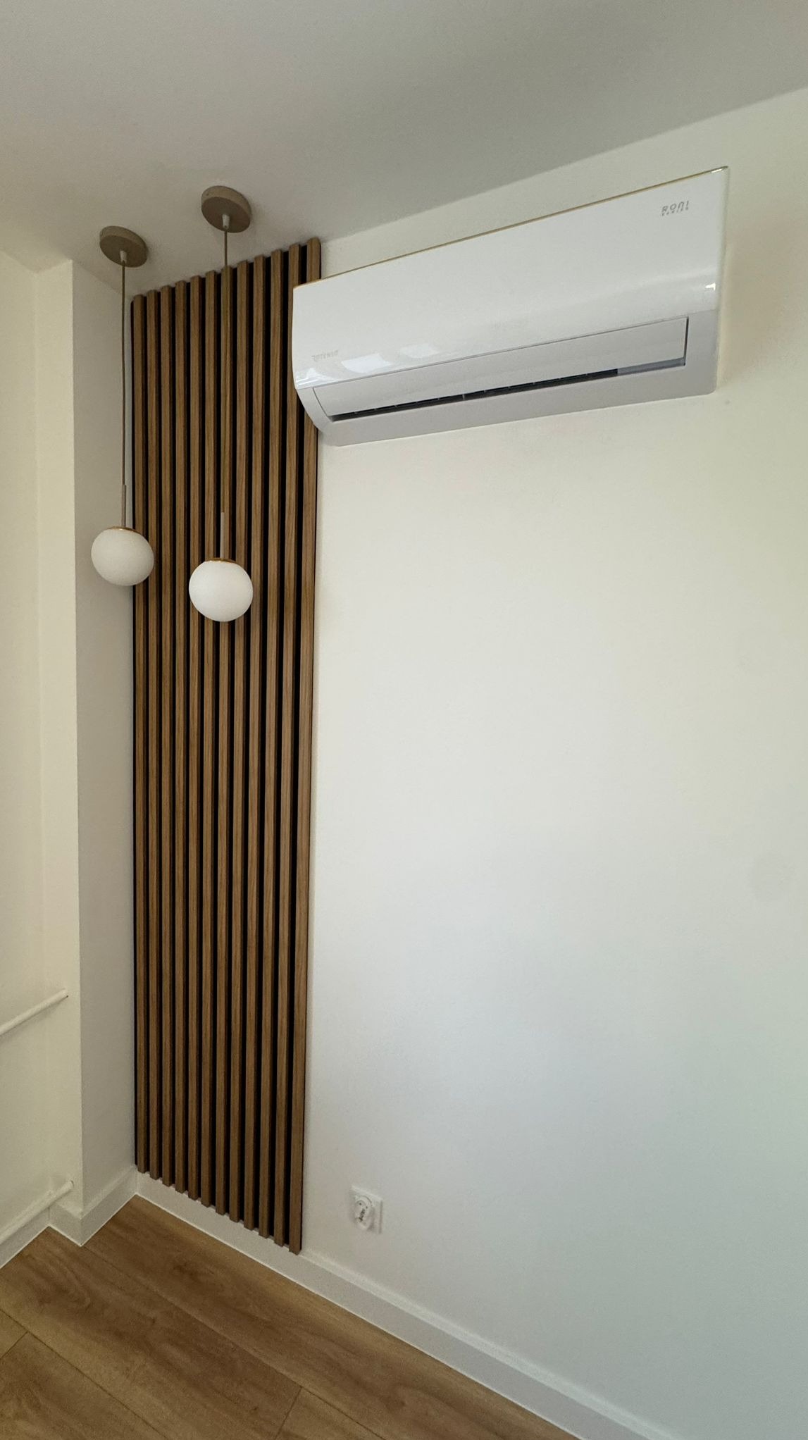 Biały klimatyzator na ścianie, obok dekoracyjna drewniana ścianka z lampami. Minimalistyczne wnętrze z drewnianą podłogą i gładkimi ścianami.