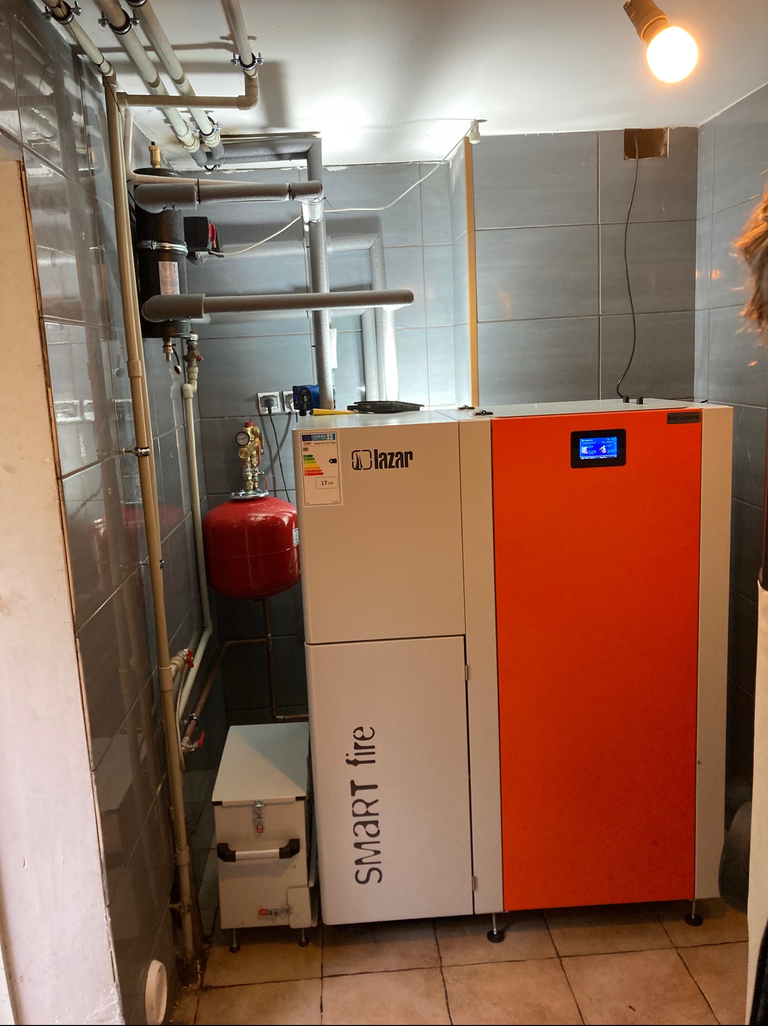 Nowoczesny piec Lazar Smart Fire z pomarańczowym panelem, zainstalowany w kotłowni z szarymi płytkami. Widoczne rury instalacji i zbiornik wyrównawczy.