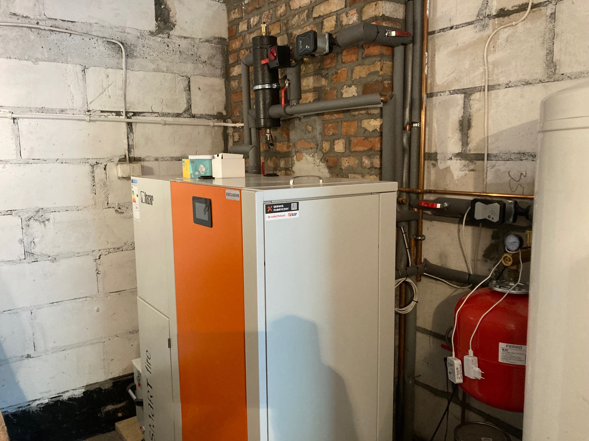 Nowoczesny piec gazowy Lazar z pomarańczowym panelem, zainstalowany w kotłowni z widocznymi rurami i elementami instalacji grzewczej na tle ściany z cegły i bloczków.