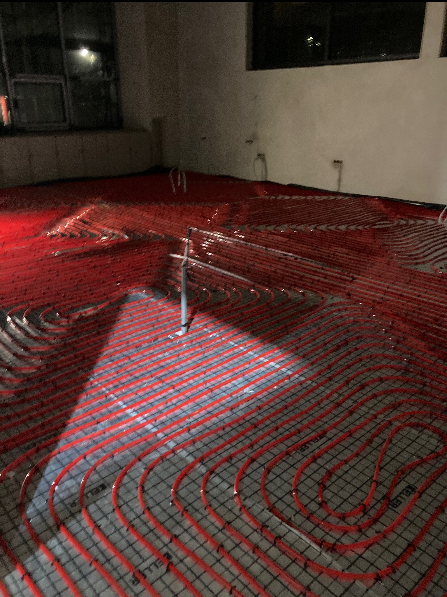 Czerwone rury ogrzewania podłogowego ułożone na siatce, widoczne elementy instalacji i fragment okna w tle. Etap prac przed wylaniem posadzki.