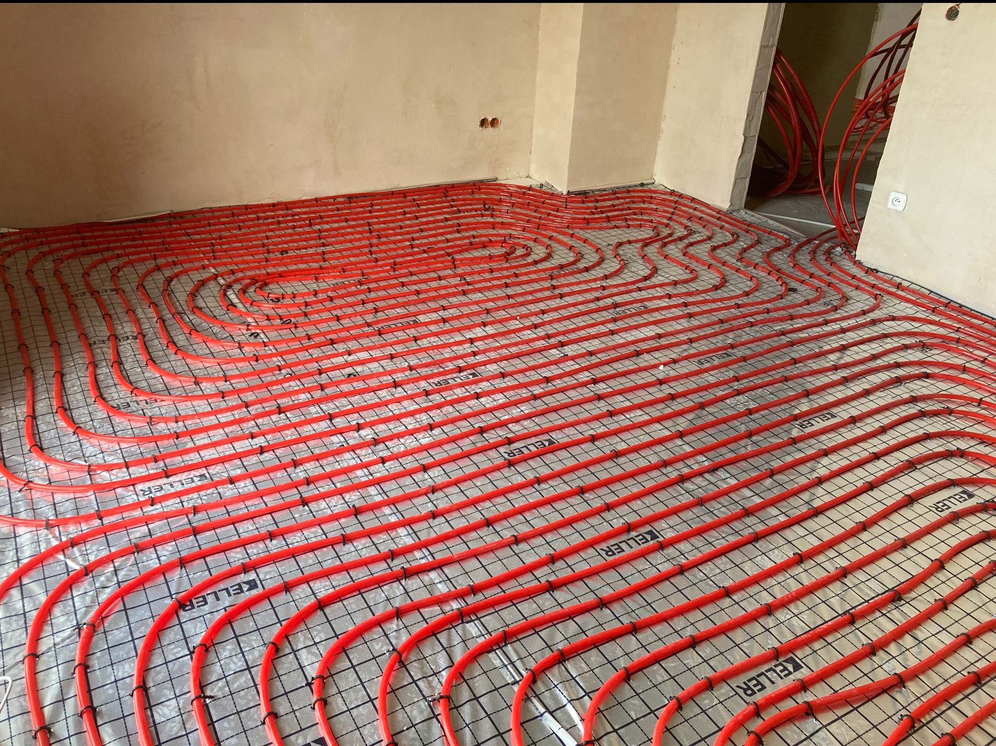 Czerwone rury ogrzewania podłogowego ułożone spiralnie na folii z siatką w surowym wnętrzu. Widoczne uchwyty mocujące rury. Etap instalacji przed wylewką.