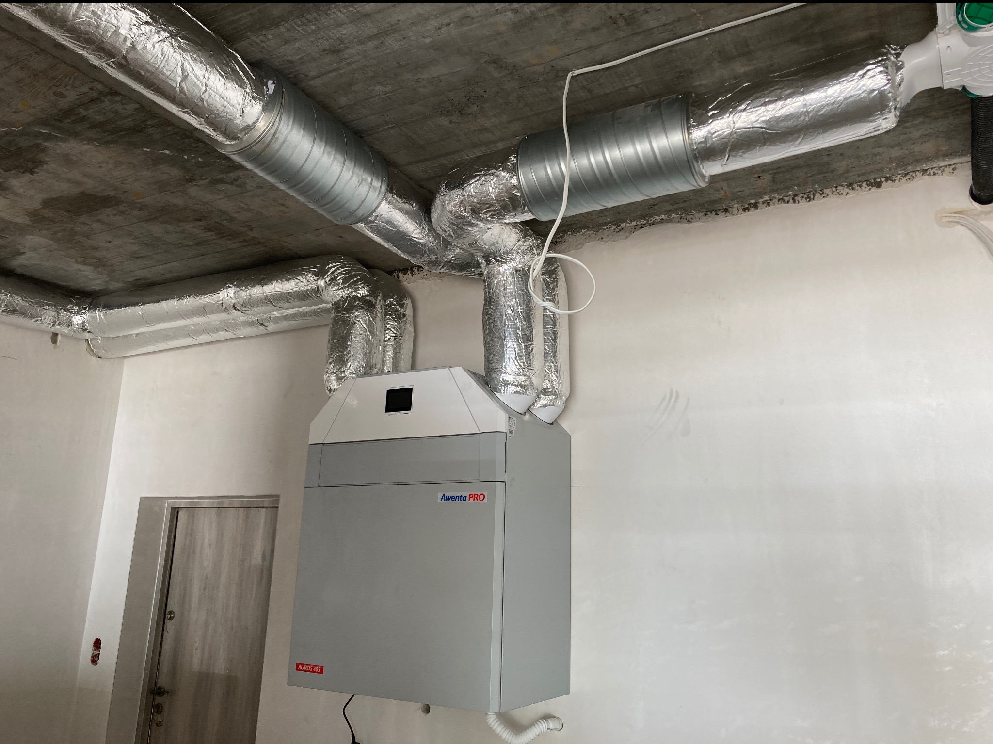 Szara centrala wentylacyjna Aventa PRO z podłączonymi, zaizolowanymi przewodami wentylacyjnymi pod betonowym stropem. Widoczny fragment ściany i drzwi.