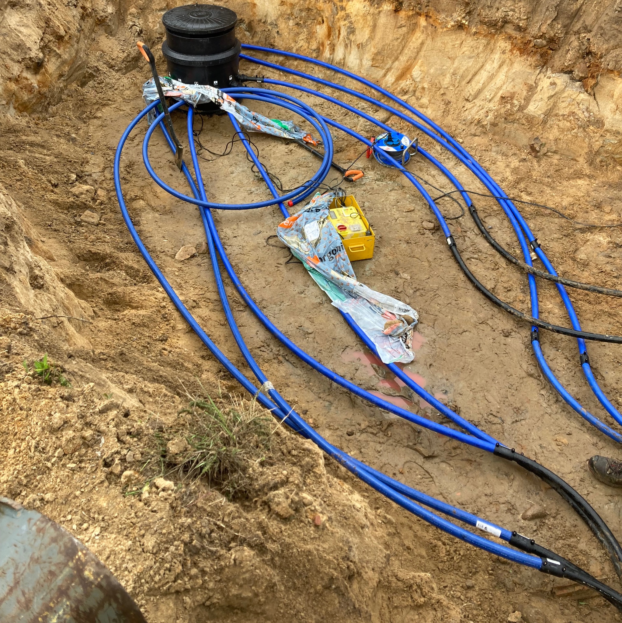Niebieskie rury ułożone w wykopie ziemnym, prowadzące do czarnej studzienki. Widoczne narzędzia i akcesoria instalacyjne. Prace ziemne w toku.