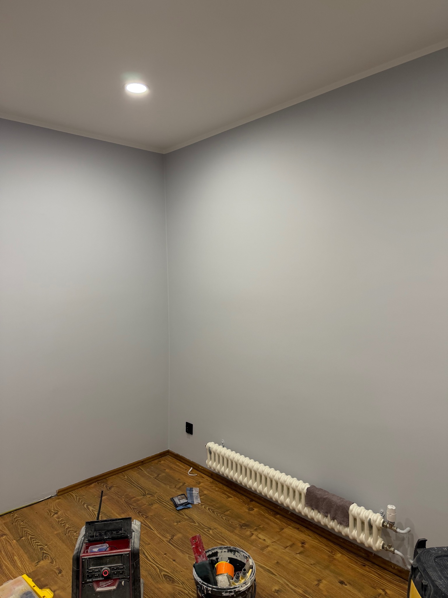 Świeżo pomalowane na szaro ściany w pokoju z drewnianą podłogą. Widoczny biały grzejnik i akcesoria malarskie: wiadro z farbą, radio budowlane.