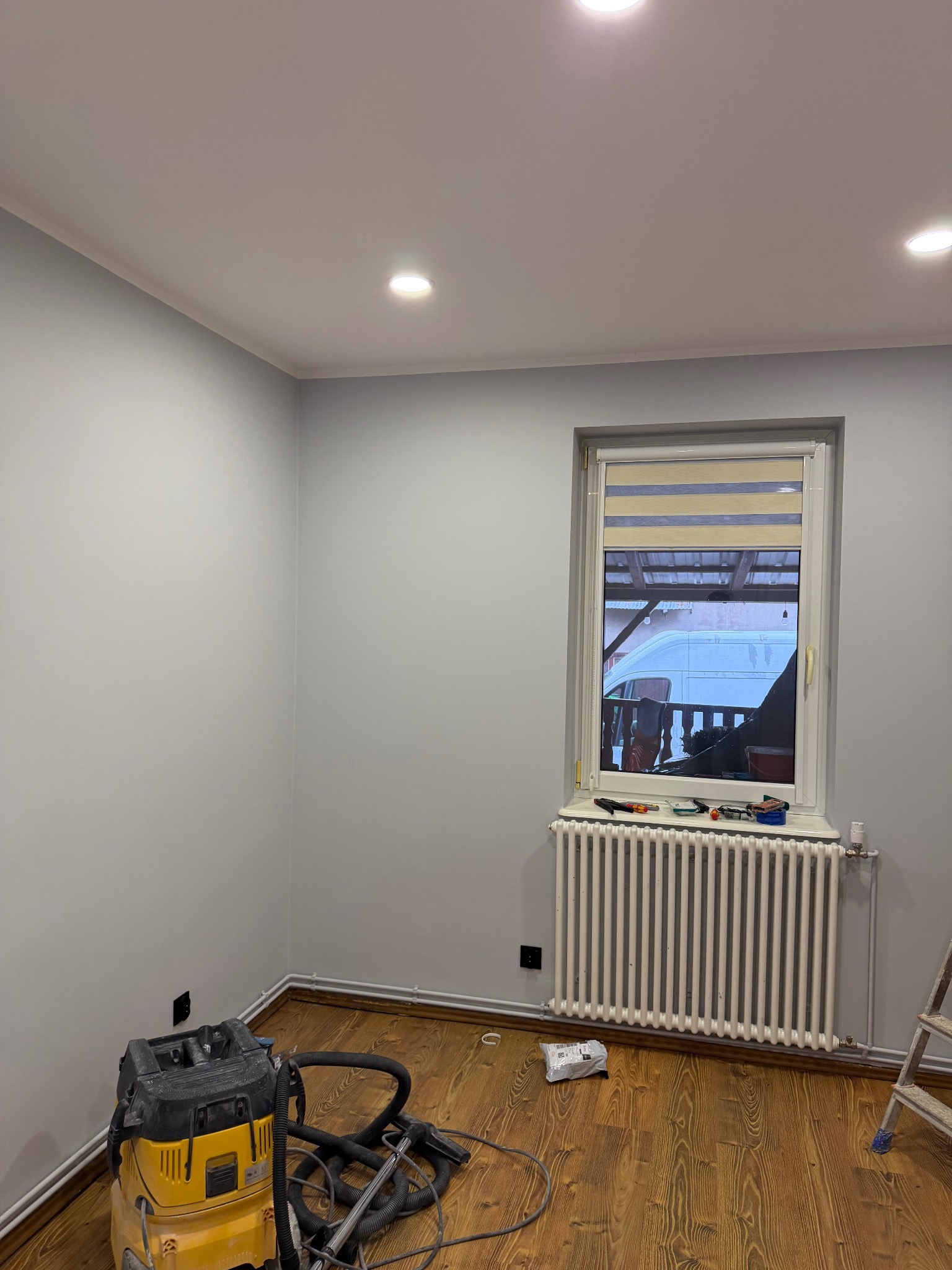 Pomieszczenie po malowaniu ścian na szaro, widoczny grzejnik, okno z roletą, odkurzacz przemysłowy i drewniana podłoga. Świeżo odmalowane ściany.