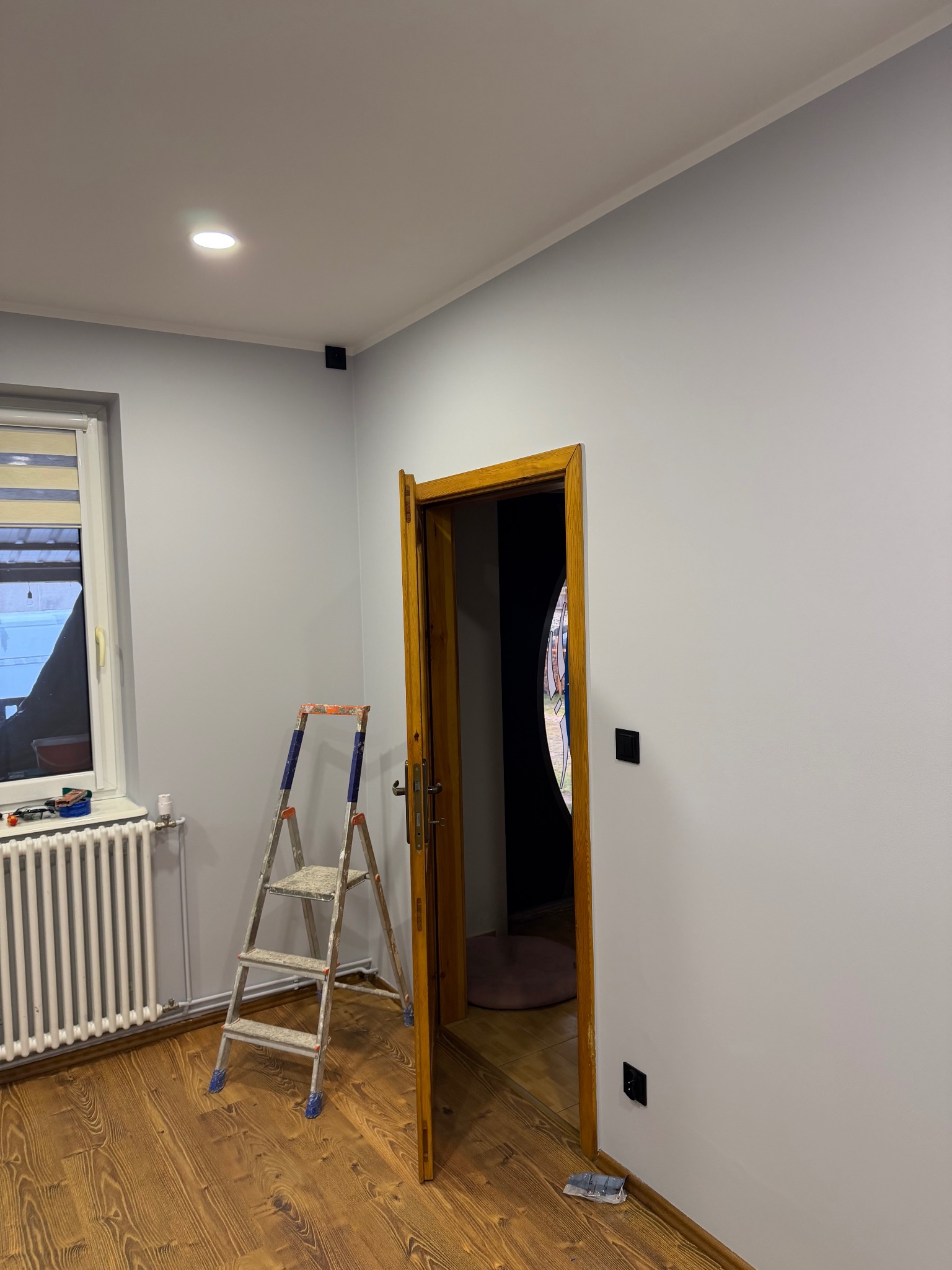 Jasnoszare ściany po malowaniu, drewniana podłoga, drabina malarska, okno z żaluzjami i drewniane drzwi wewnętrzne. Widoczne gniazdka elektryczne.