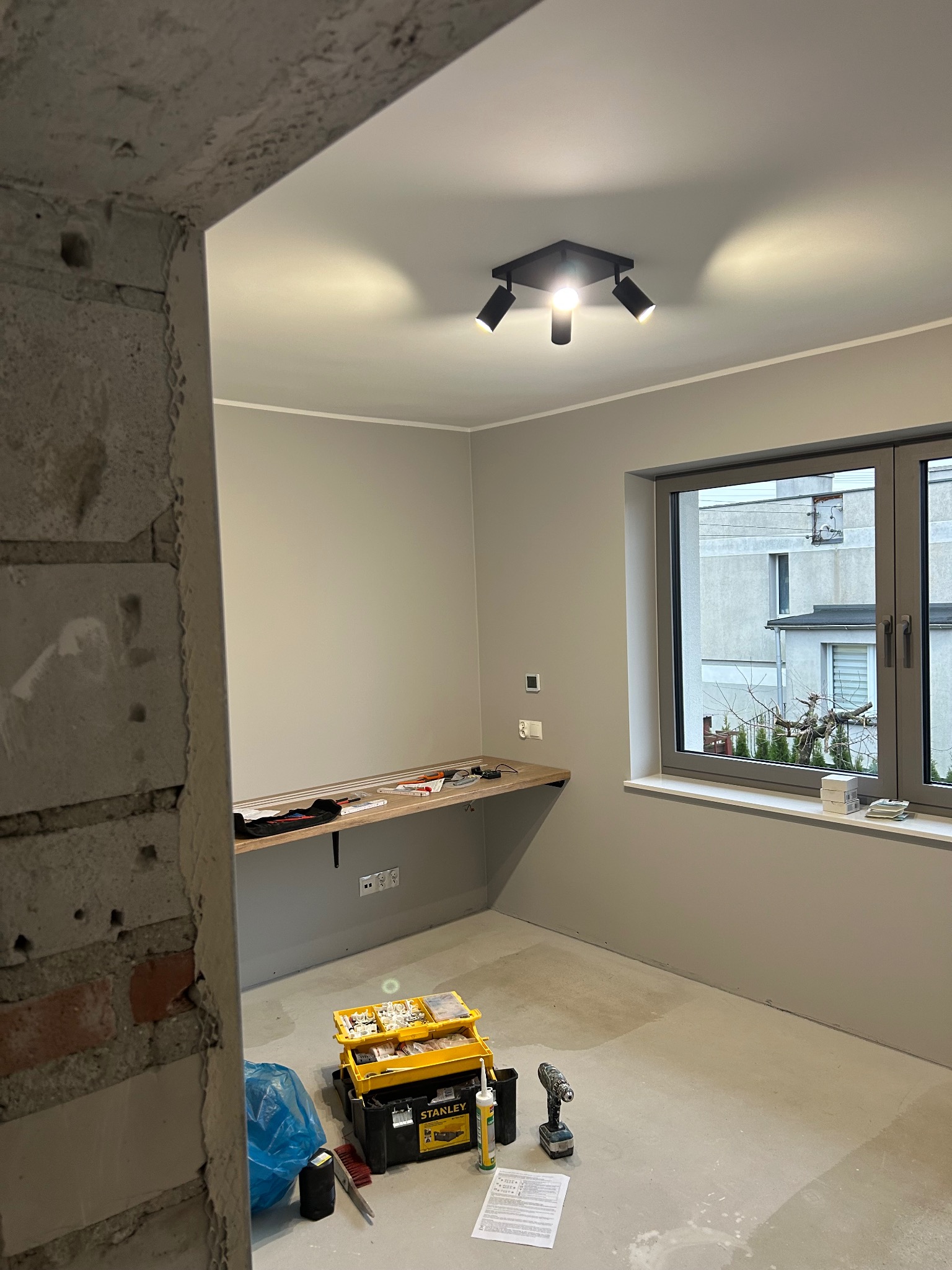 Szare wnętrze pokoju po remoncie z narzędziami Stanley, lampą sufitową i oknem. Widoczny fragment surowej ściany z cegły.