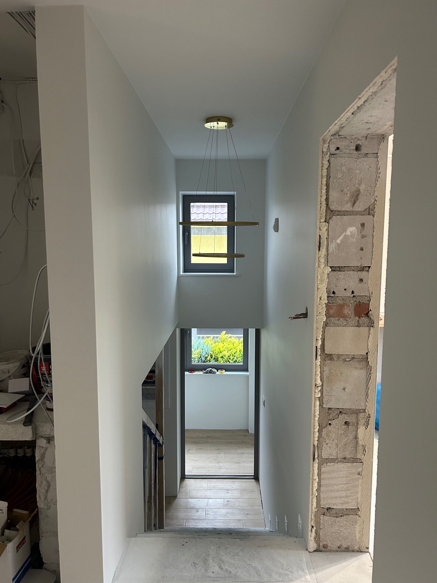 Korytarz w trakcie remontu: świeżo pomalowane ściany w odcieniach bieli, widoczne okna, schody i surowa ściana z cegieł, nowoczesna lampa wisząca.