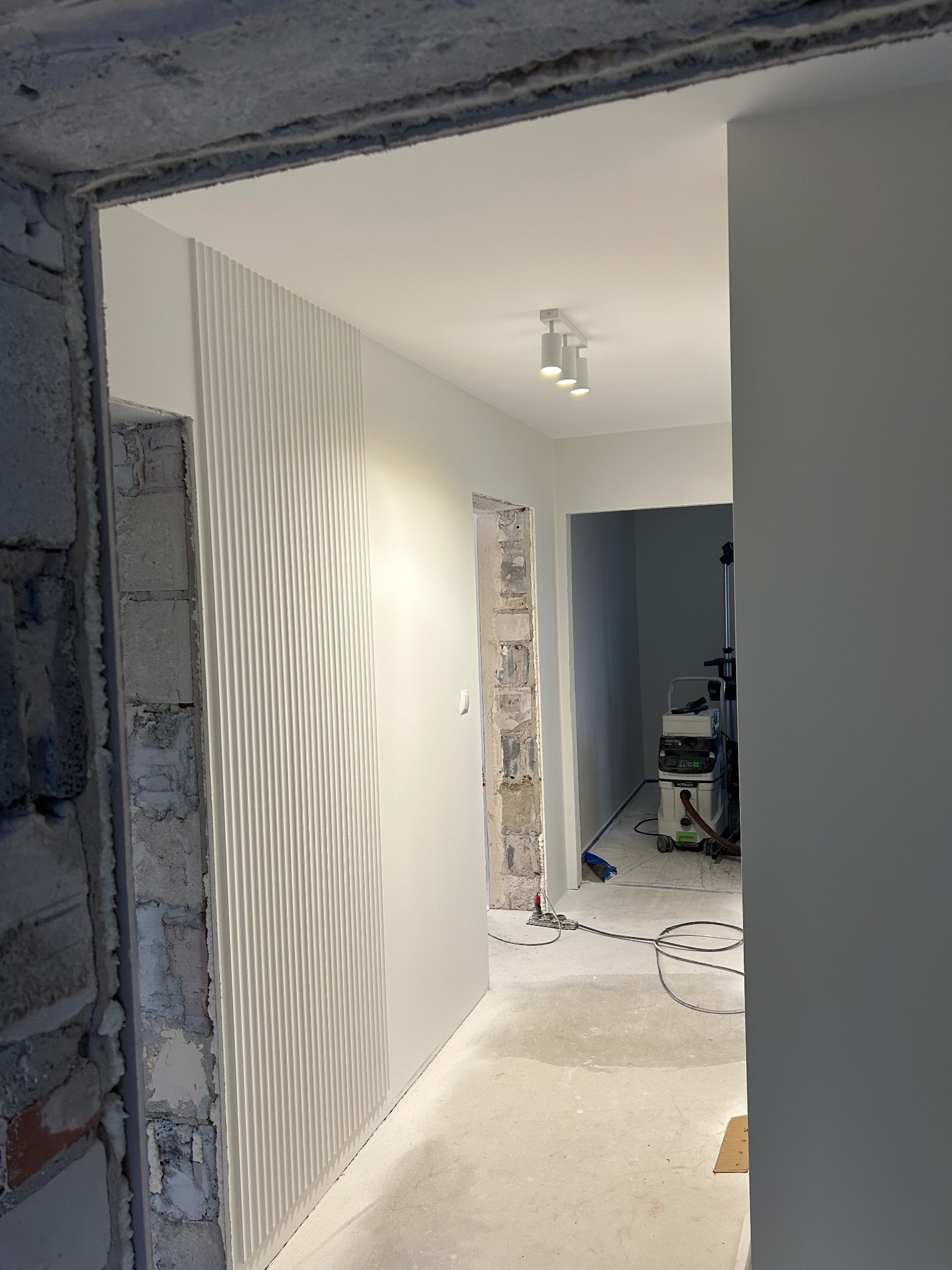 Białe lamele na ścianie w korytarzu w trakcie remontu. Widoczne surowe ściany i sprzęt budowlany. Oświetlenie sufitowe punktowe.