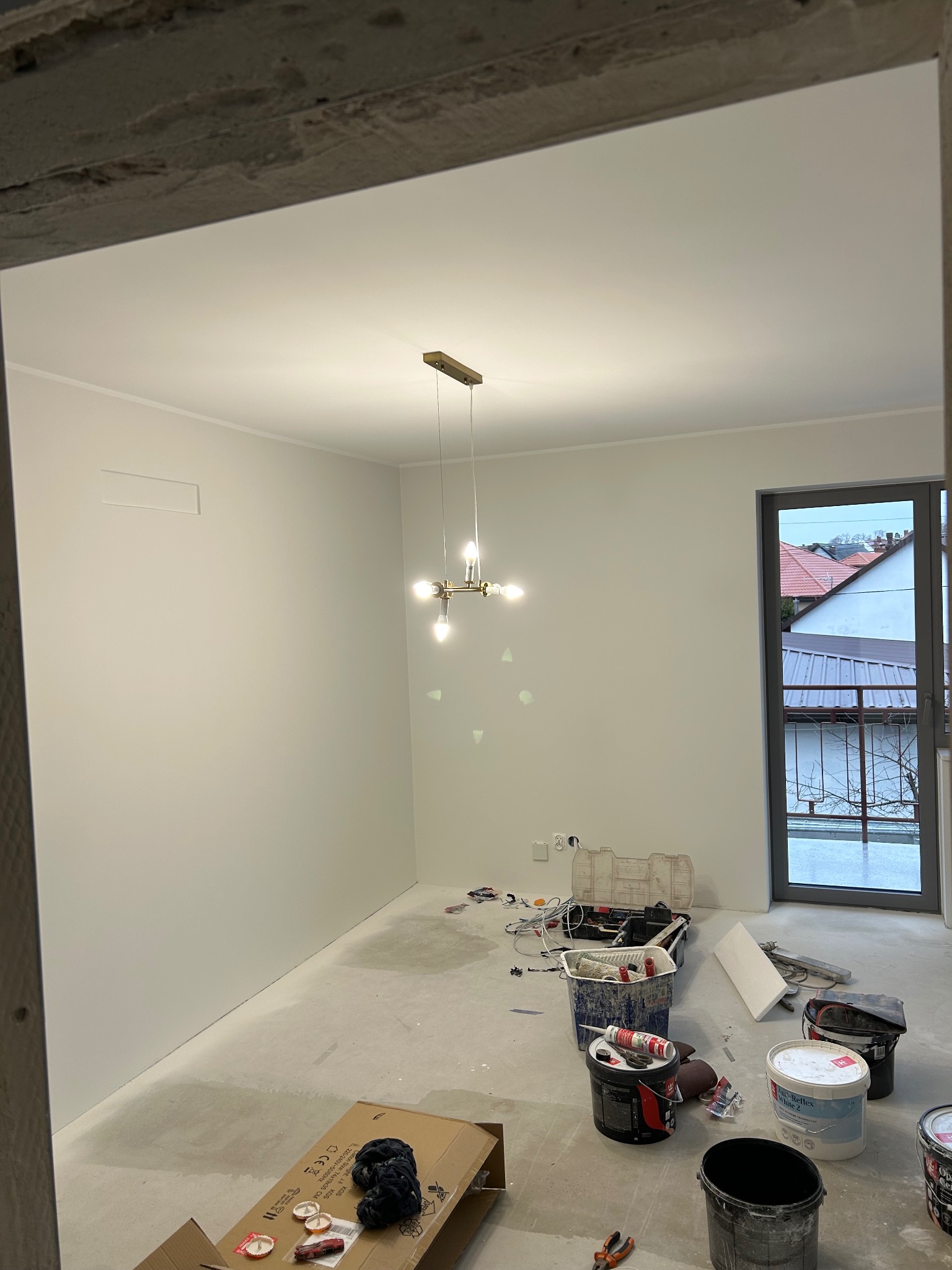 Wnętrze po malowaniu ścian na biało, widoczne narzędzia malarskie, pędzle, wiadra z farbą, kartony. Nowoczesna lampa sufitowa. Okno z widokiem na domy.
