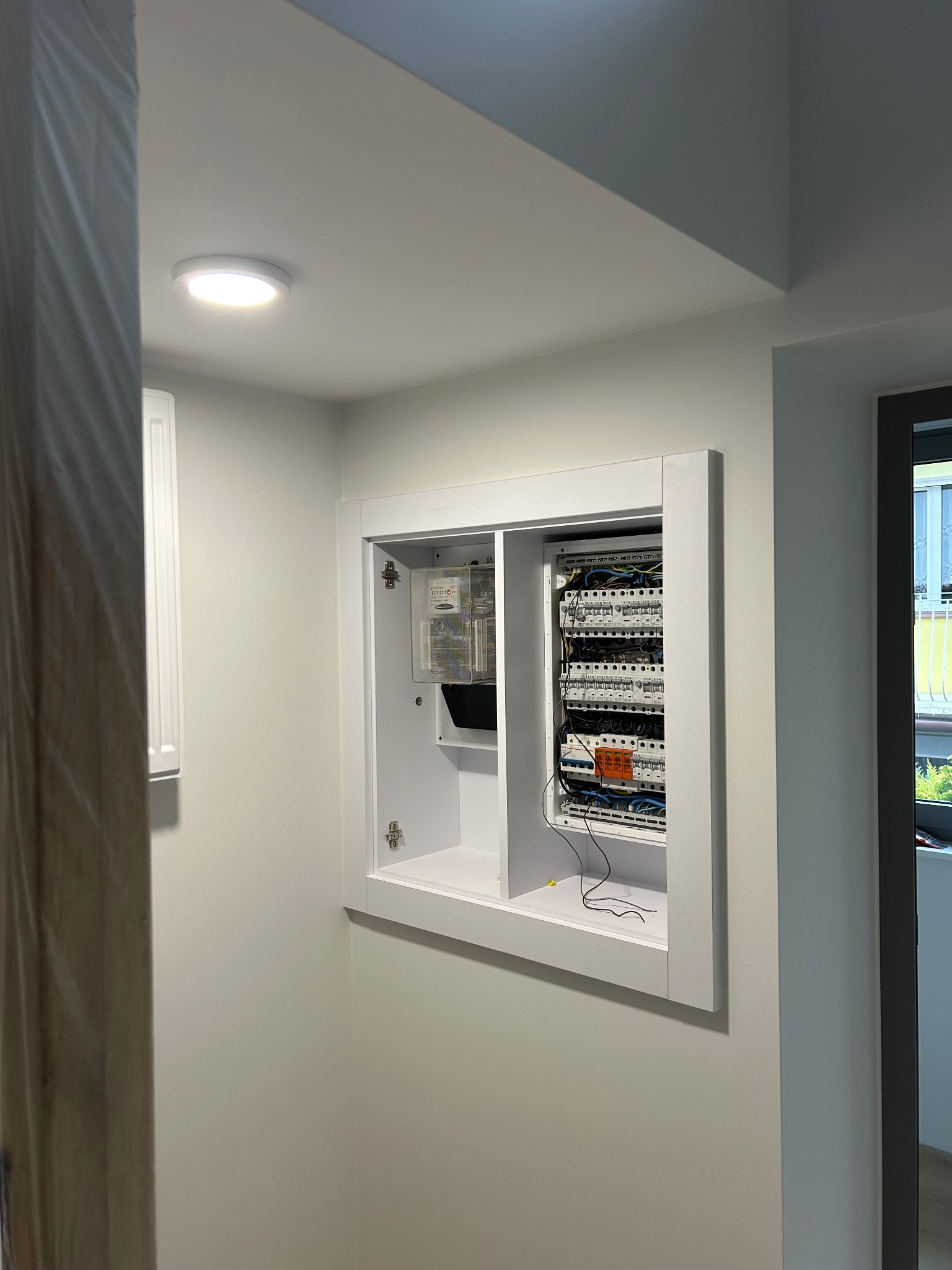 Wnęka z białą obudową kryjąca rozdzielnicę elektryczną i licznik, widoczne przewody. Minimalistyczne wnętrze z oświetleniem punktowym.
