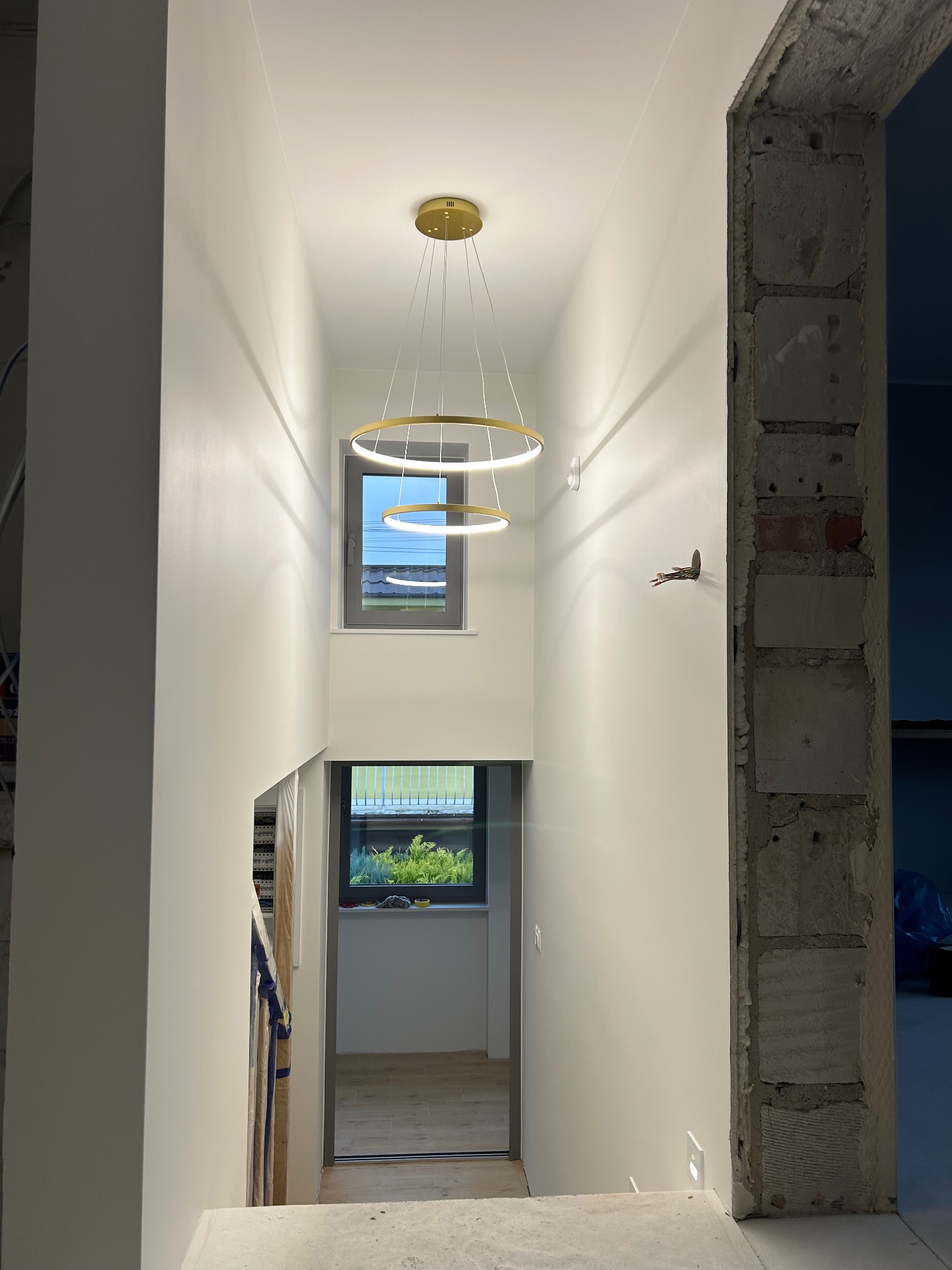 Wnętrze po malowaniu w Bysławiu: białe ściany, nowoczesna lampa wisząca, widok na okno i schody. Fragment surowego muru przy przejściu.