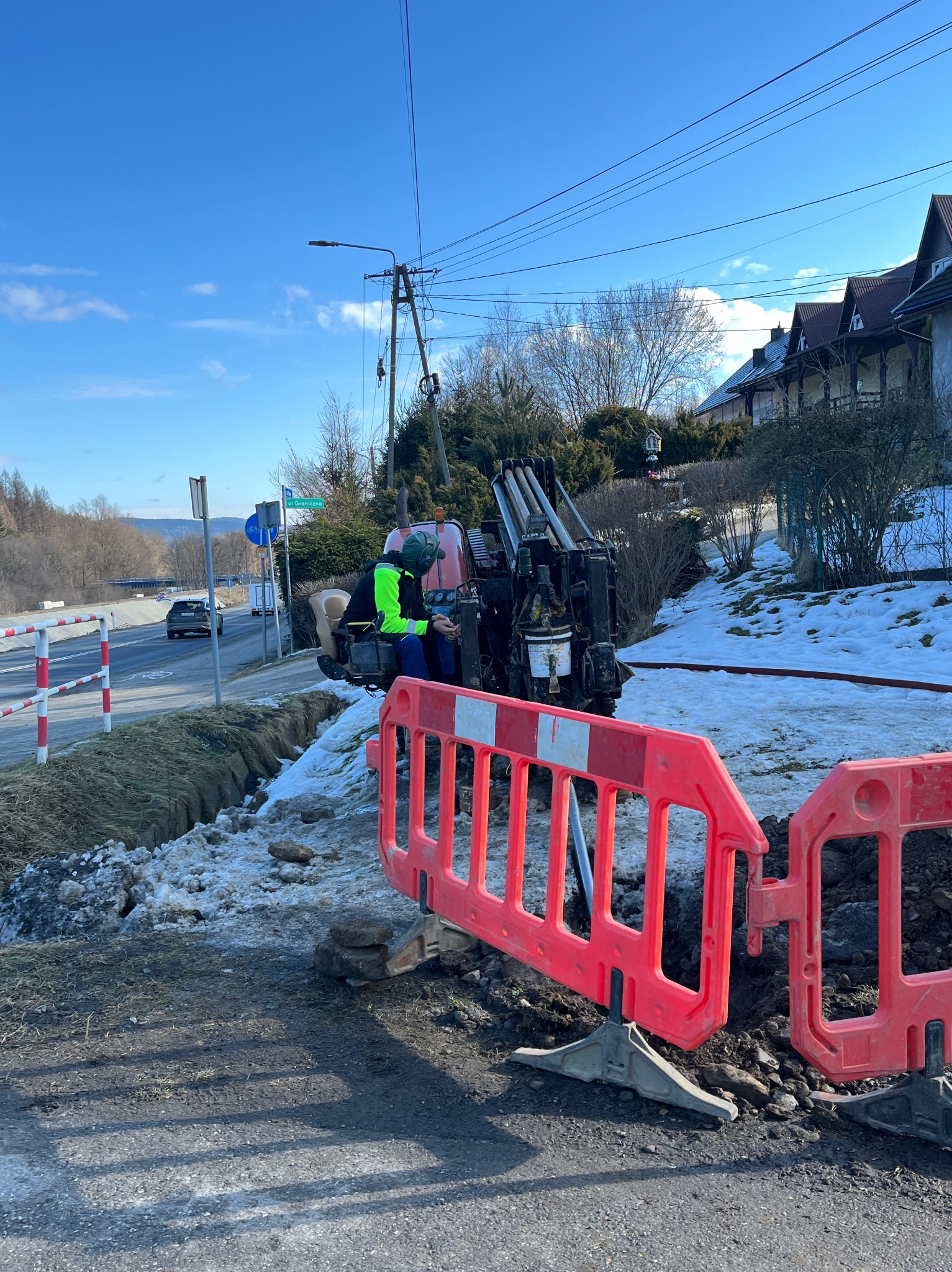 Operator w neonowej kamizelce obsługuje maszynę do robót ziemnych na tle krajobrazu z drogą i zabudowaniami. Widoczne czerwone bariery ochronne.