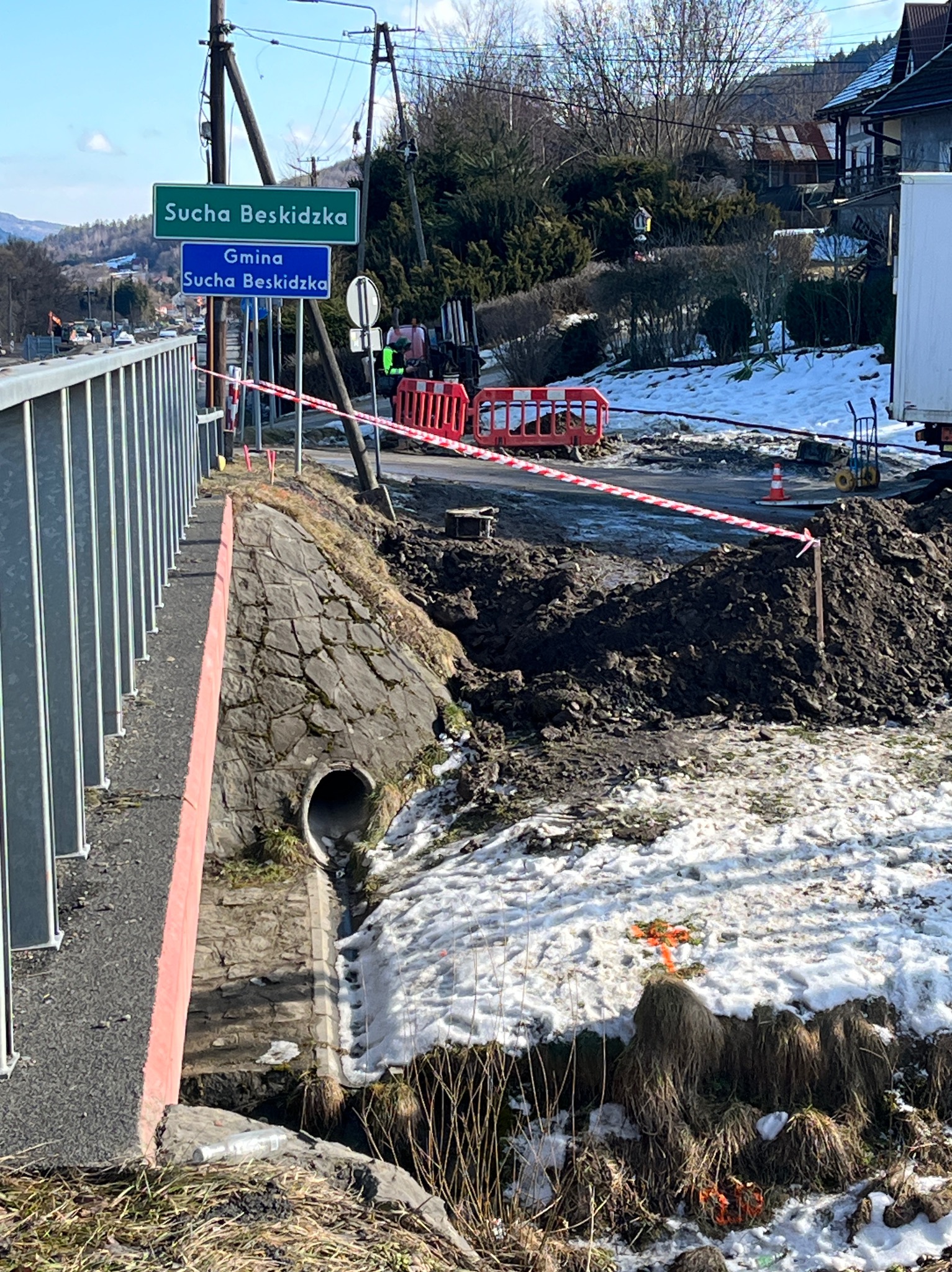 Wykonanie przepustu pod drogą w terenie górzystym, zabezpieczone barierami i taśmami ostrzegawczymi. Widoczne znaki drogowe z nazwą miejscowości.