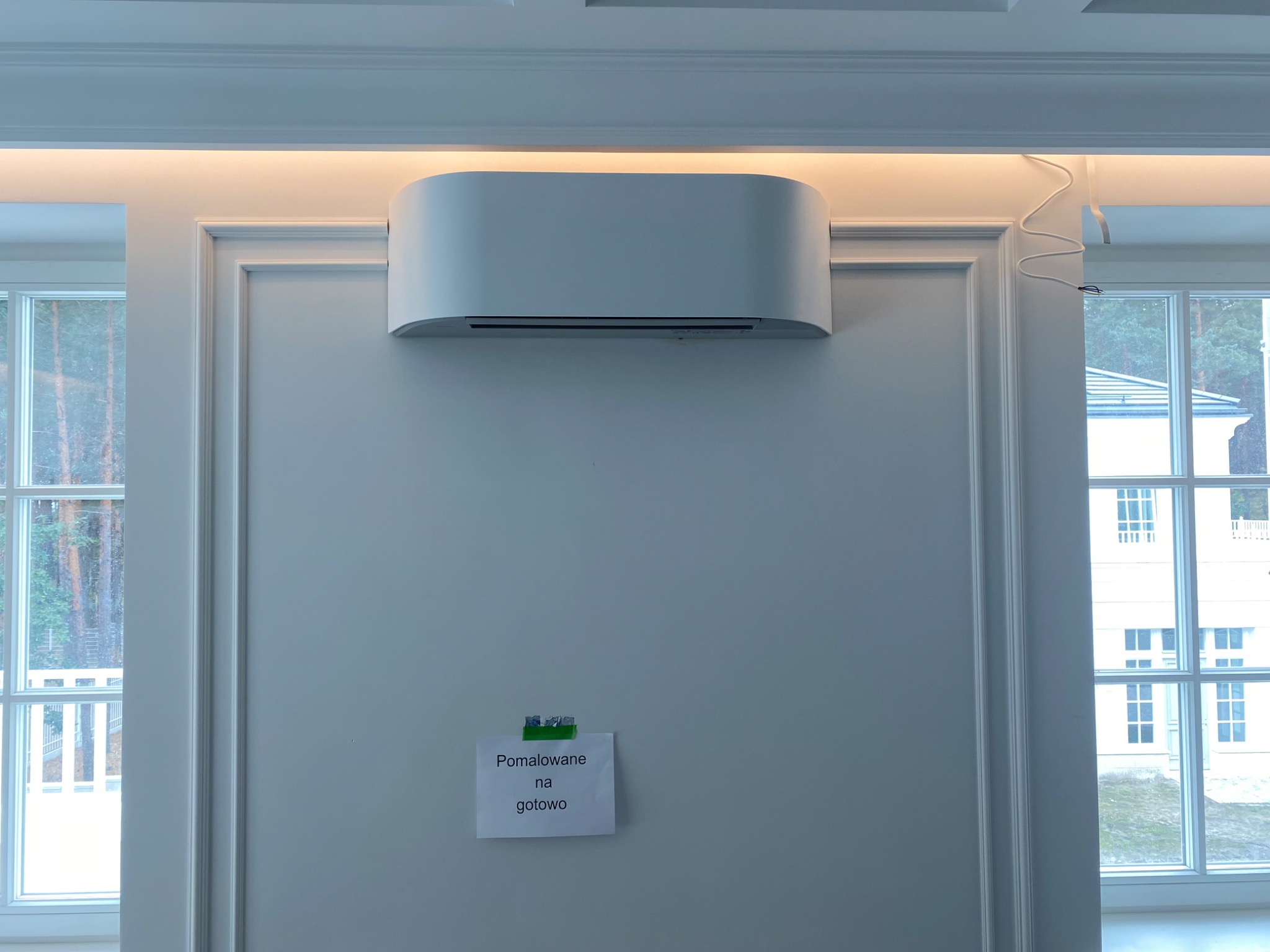 Nowoczesny, biały klimatyzator ścienny w eleganckim wnętrzu z listwami dekoracyjnymi i oknami. Widoczna kartka z napisem 'Pomalowane na gotowo'.