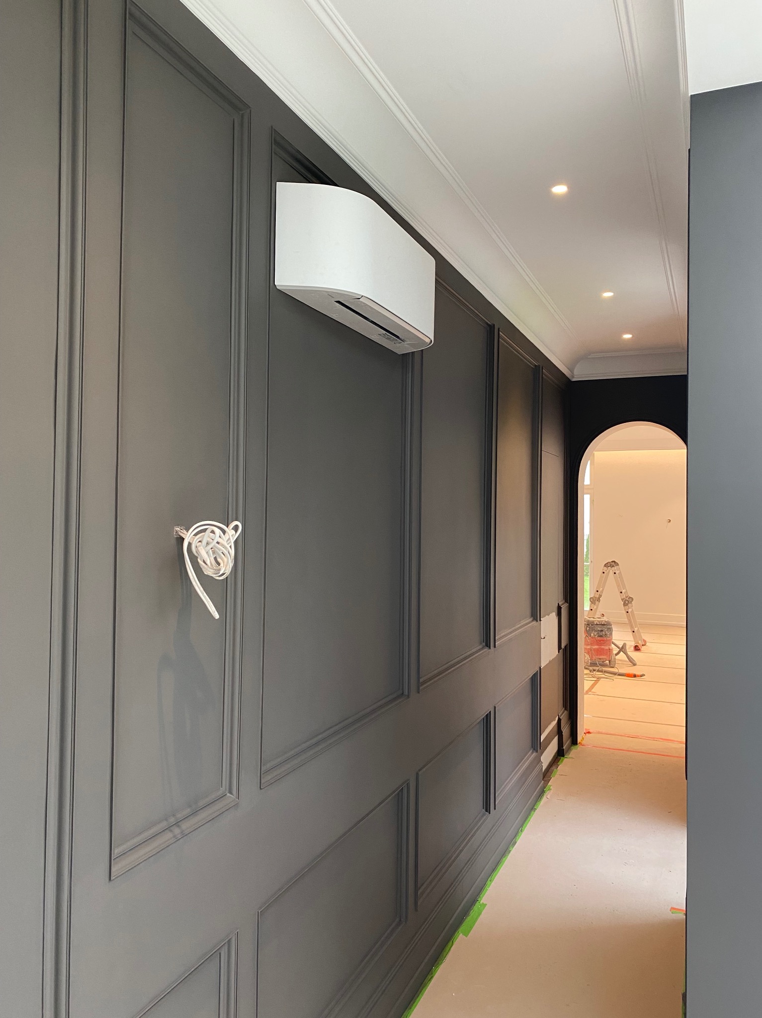 Biała klimatyzacja ścienna w eleganckim, szarym wnętrzu z panelami. Widoczne przewody i drabina w tle sugerują etap instalacji lub serwisu.