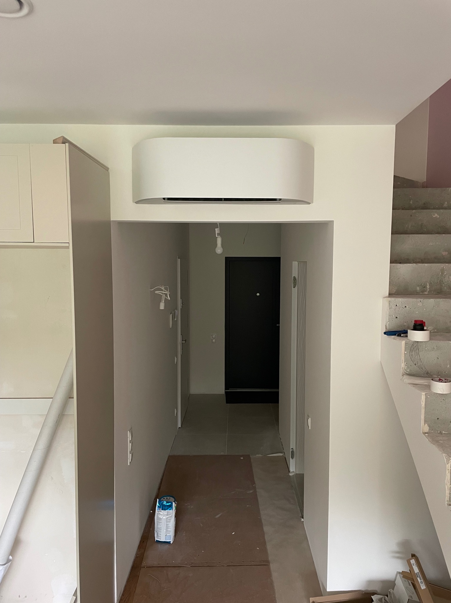 Biała, zaokrąglona jednostka klimatyzacyjna zamontowana nad wejściem do korytarza w jasnym wnętrzu. Widoczne schody w trakcie wykończenia i materiały budowlane.