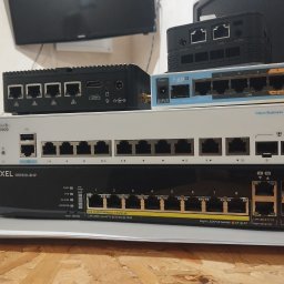 SYSNET-ELECTRO.COM Artur Gęgotek - Stos routerów i switchy sieciowe różnych marek (Cisco, Zyxel, MikroTik) ułożone jeden na drugim na drewnianym blacie. Widoczne porty Ethernet i oznaczenia modeli.