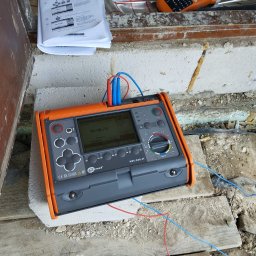 SYSNET-ELECTRO.COM Artur Gęgotek - Miernik elektryczny Sonel MPI-530-IT na budowie, wyświetlający wynik pomiaru. Widoczne kable pomiarowe, instrukcja obsługi i etykieciarka Brother.