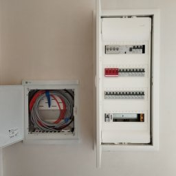 SYSNET-ELECTRO.COM Artur Gęgotek - Otwarta skrzynka elektryczna z bezpiecznikami i przewodami w ścianie. Widoczne kable ułożone w pętle w drugiej skrzynce. Białe elementy na jasnym tle ściany.